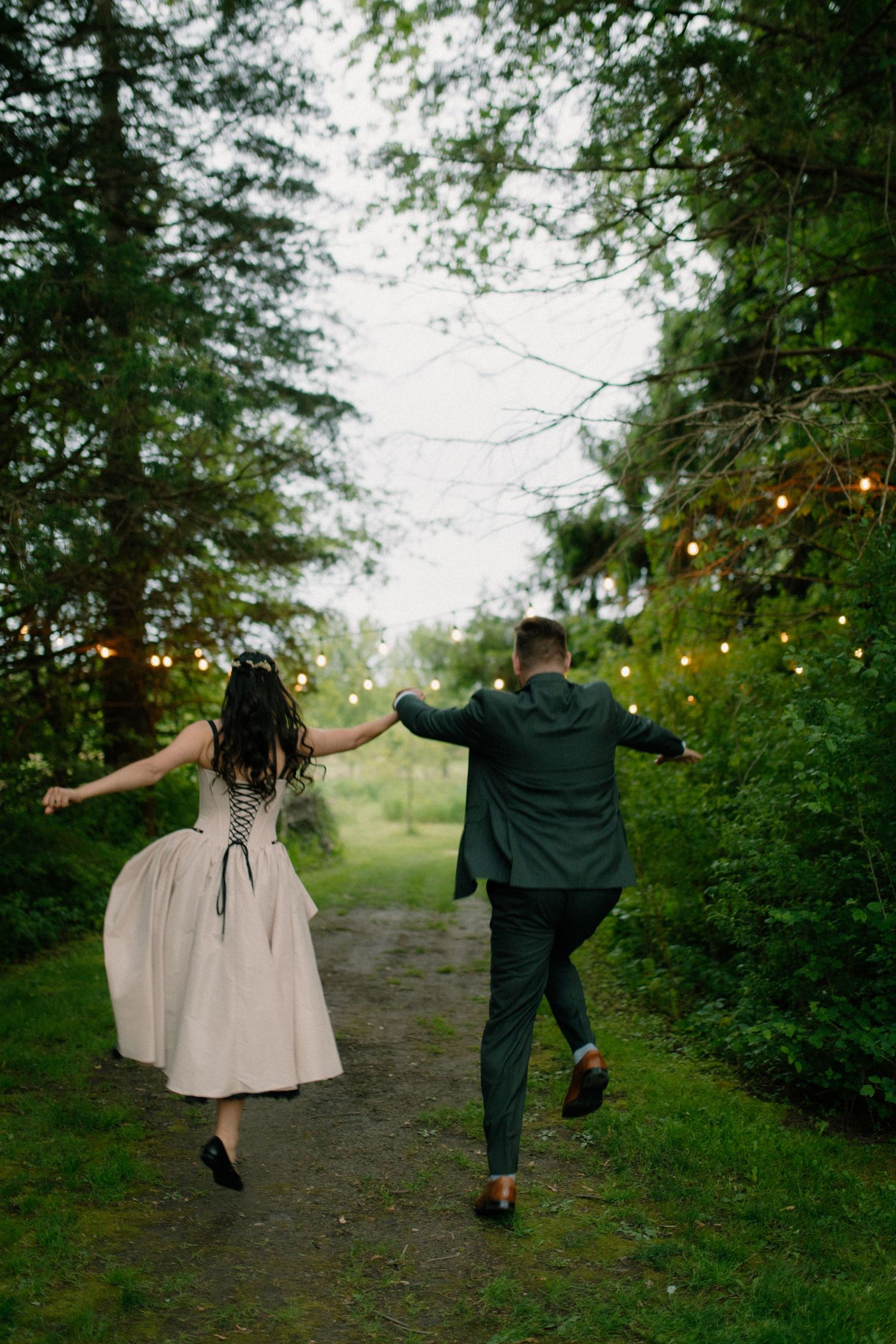 A bride and groom hold hands while joyfully running down a tree-lined path adorned with string lights.