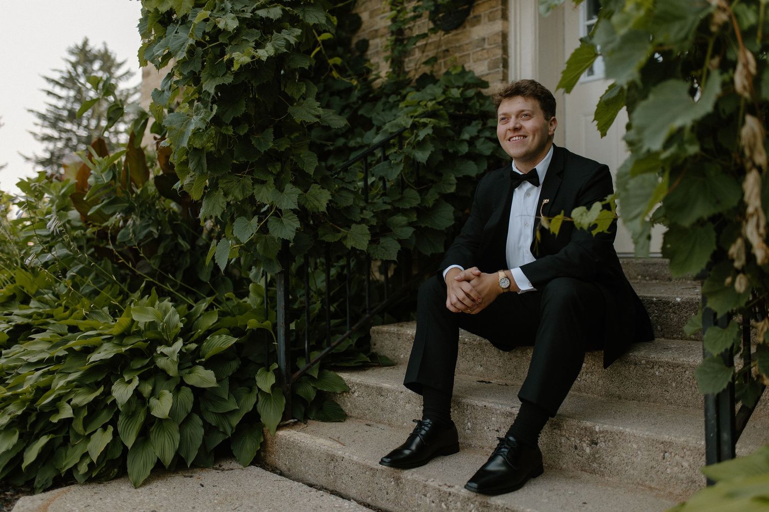 Formal portrait on ivy-covered stone steps in classic black tuxedo for Wisconsin garden wedding