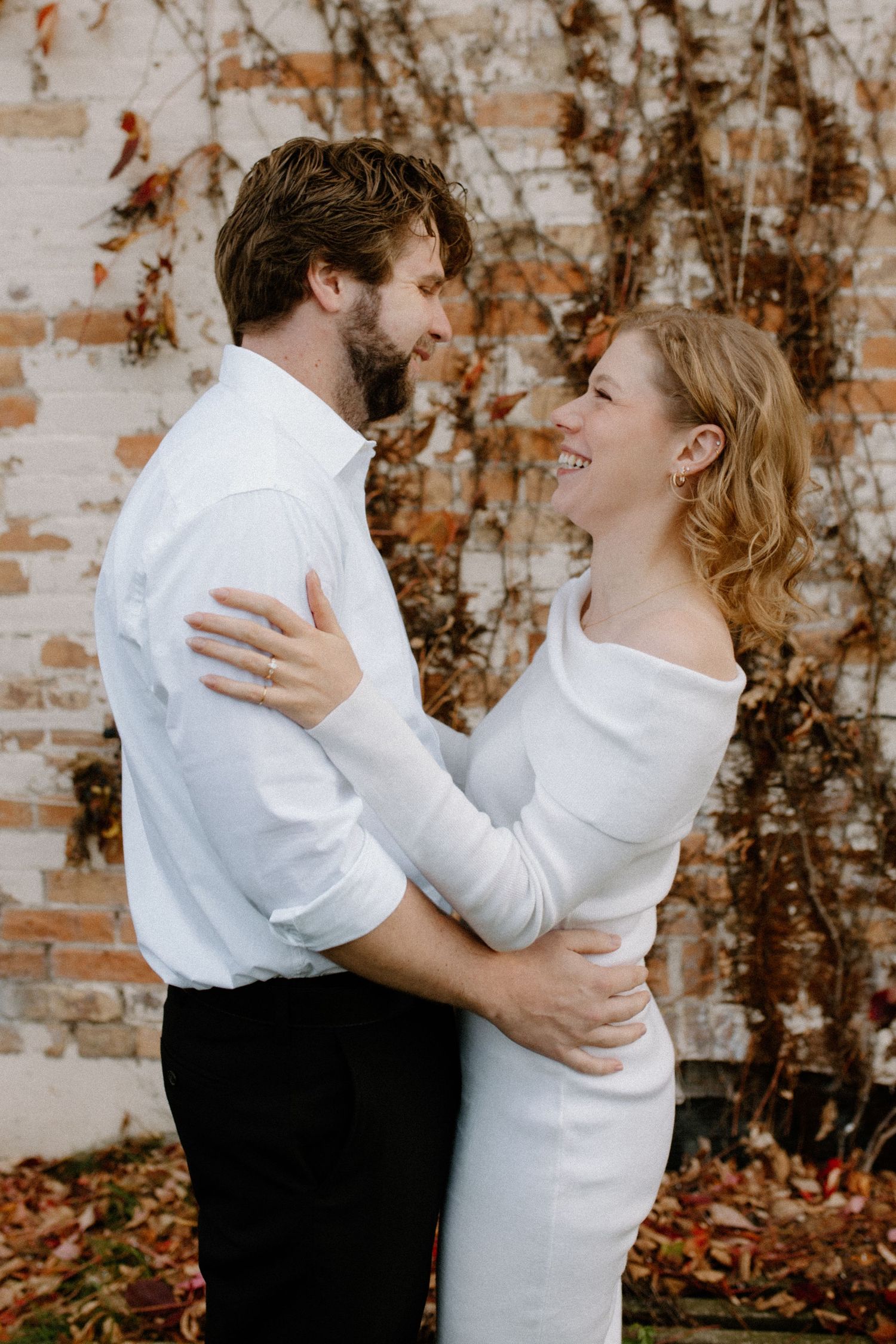 A couple in white and black attire embraces joyfully against a rustic brick wall with vines during downtown Oconomowoc engagement session