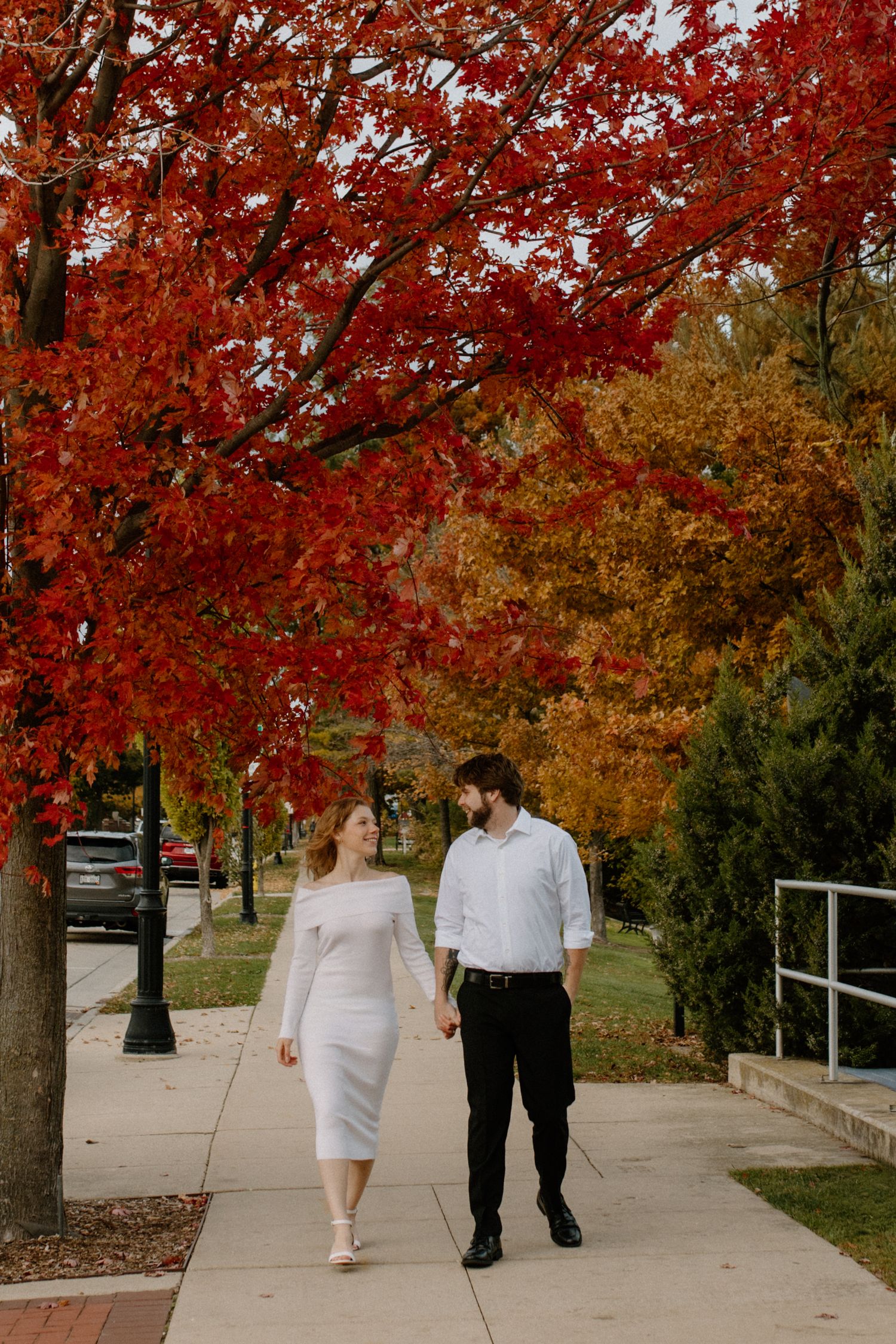 A couple strolls beneath vibrant red fall foliage on a sidewalk in downtown Oconomowoc engagement session
