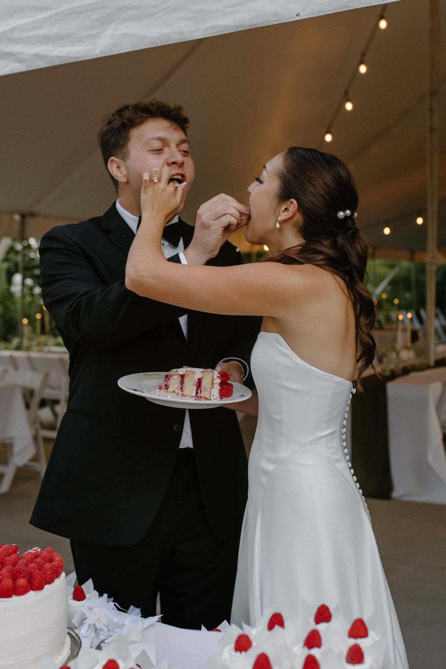 Bride and groom feeding each other wedding cake slices topped with raspberries under a tent at their reception.