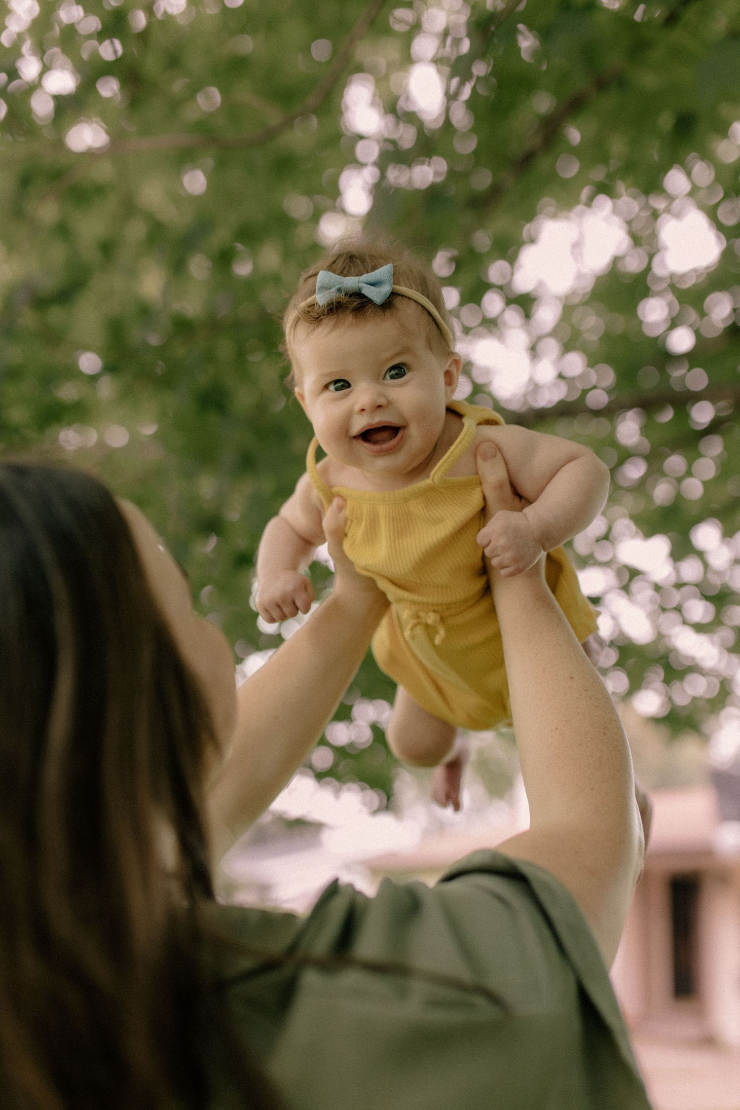 Baby wearing yellow outfit being lifted joyfully in the air against bokeh background for outdoor family photos