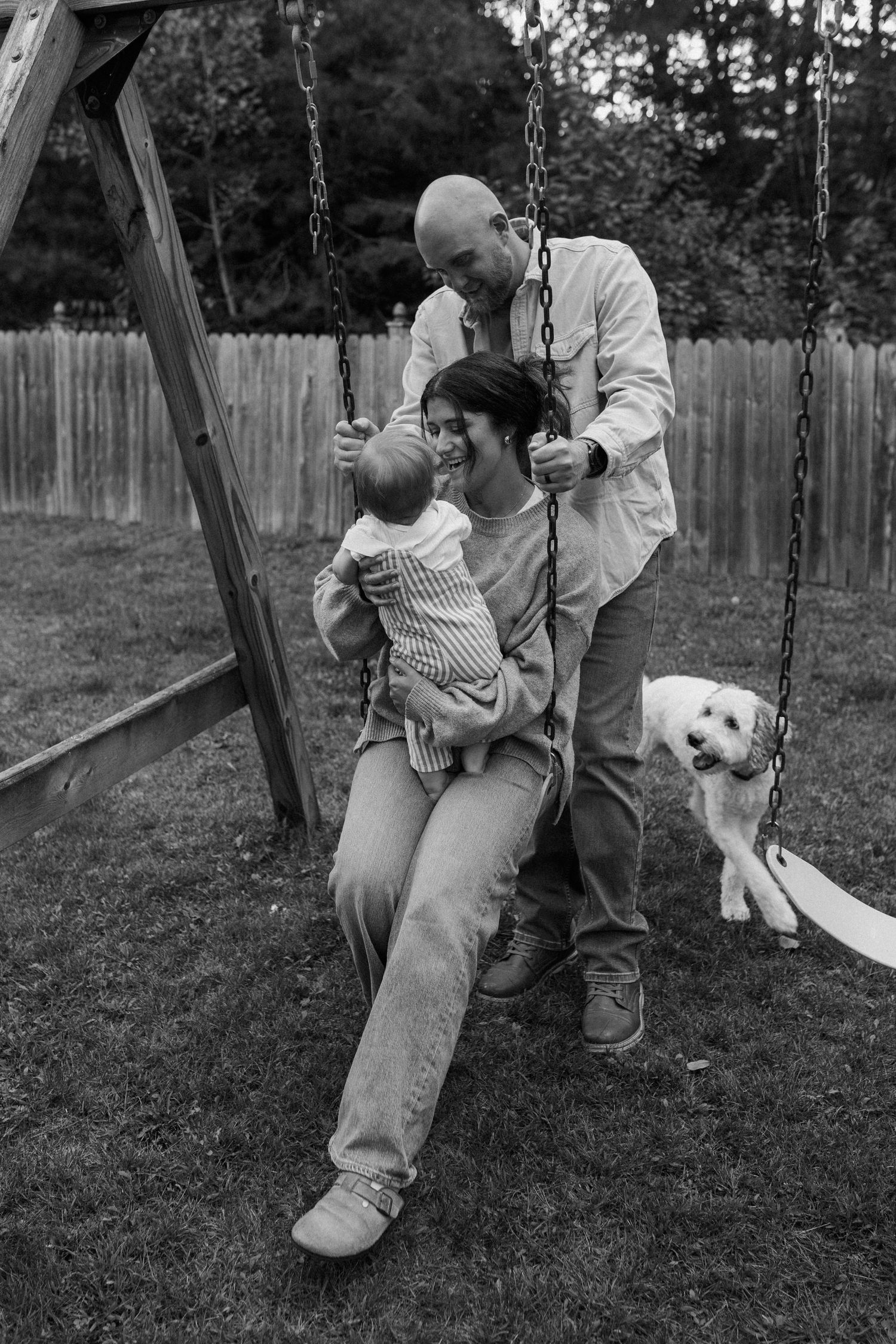 A family enjoys time together on a backyard swing set with their white dog during lifestyle photo session