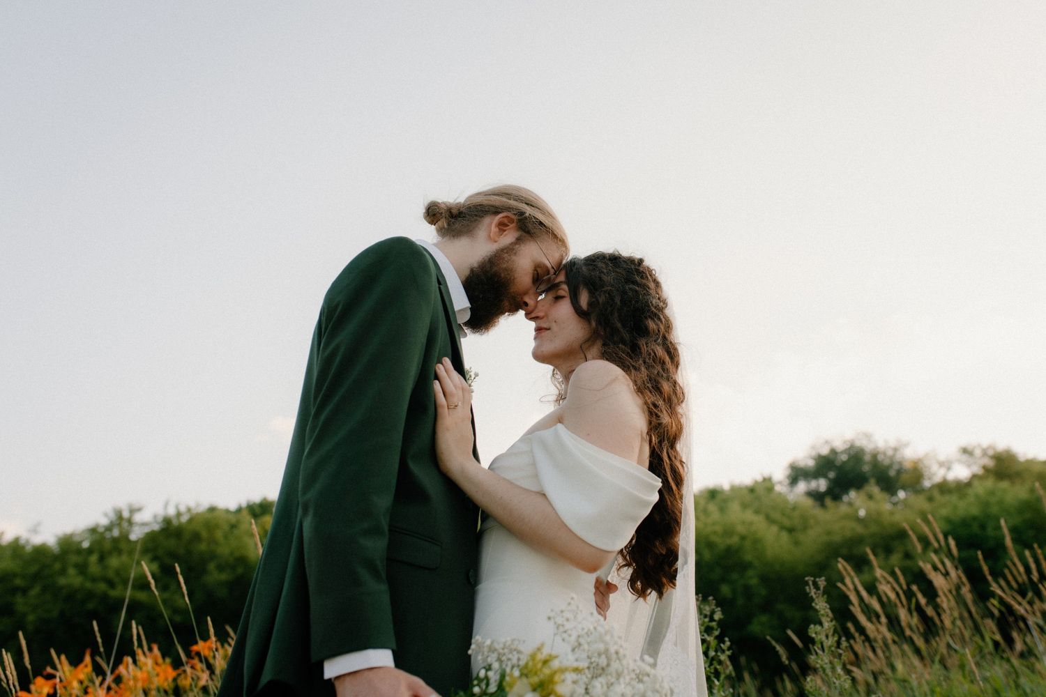A couple in wedding attire shares a romantic moment outdoors, with the groom in a green suit and bride in a white dress.