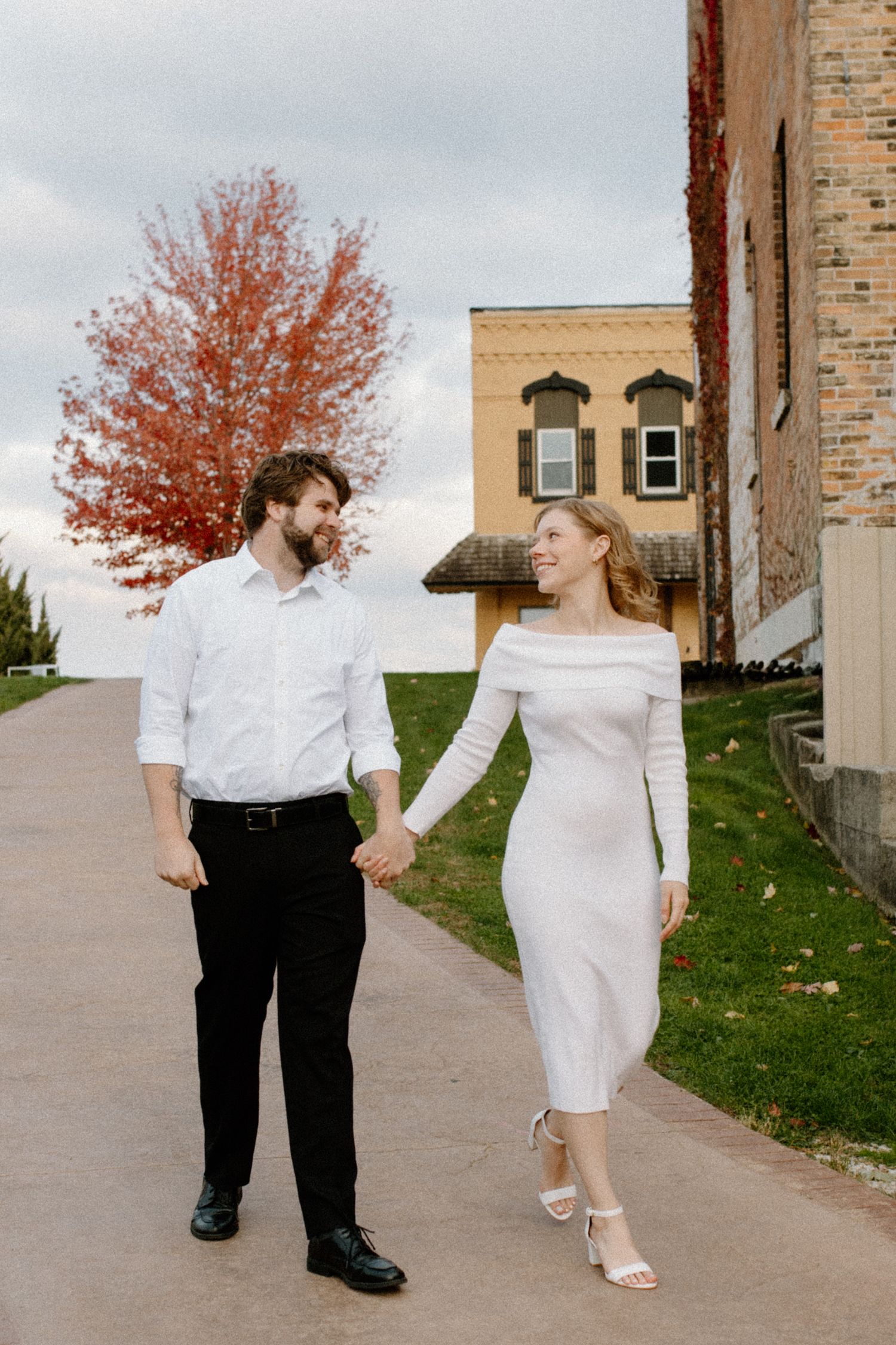 Engaged couple in formal attire walks together in downtown Oconomowoc engagement session with fall foliage in background