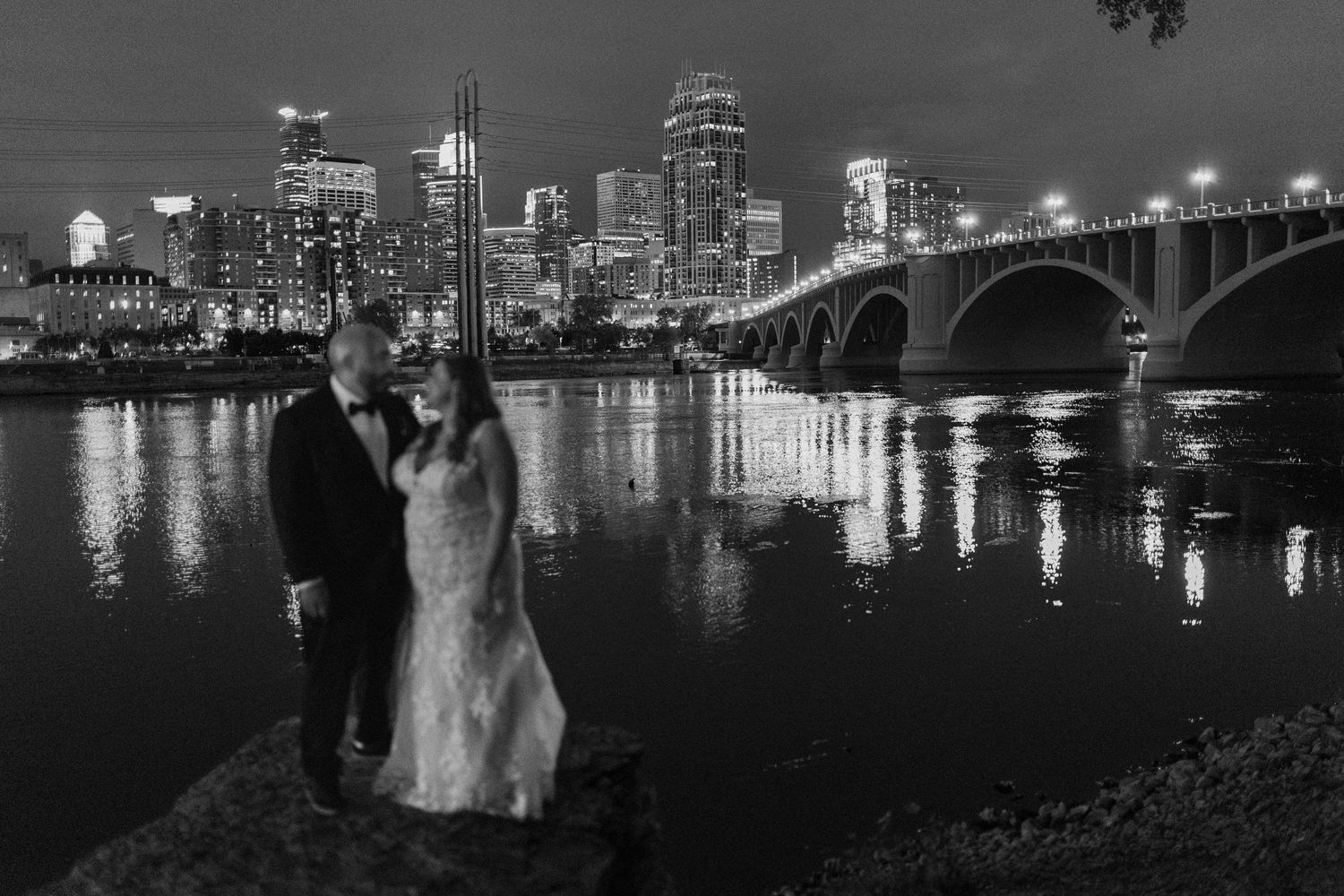 Bride and groom pose for wedding photos along Minneapolis cityscape at night with bridge and buildings reflecting on water