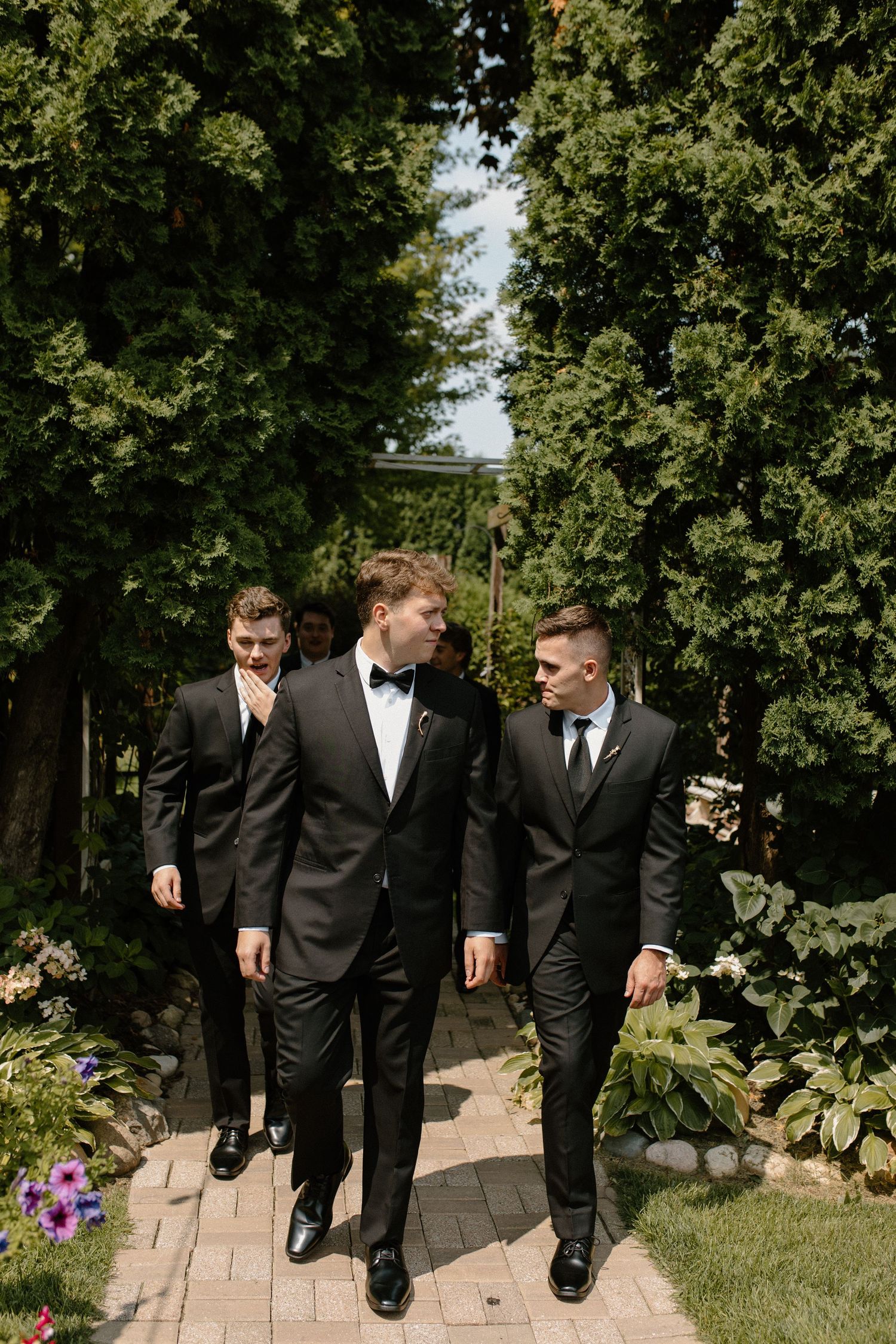 Three groomsmen in formal suits walk along a garden path lined with trees at Wisconsin Garden Wedding Center