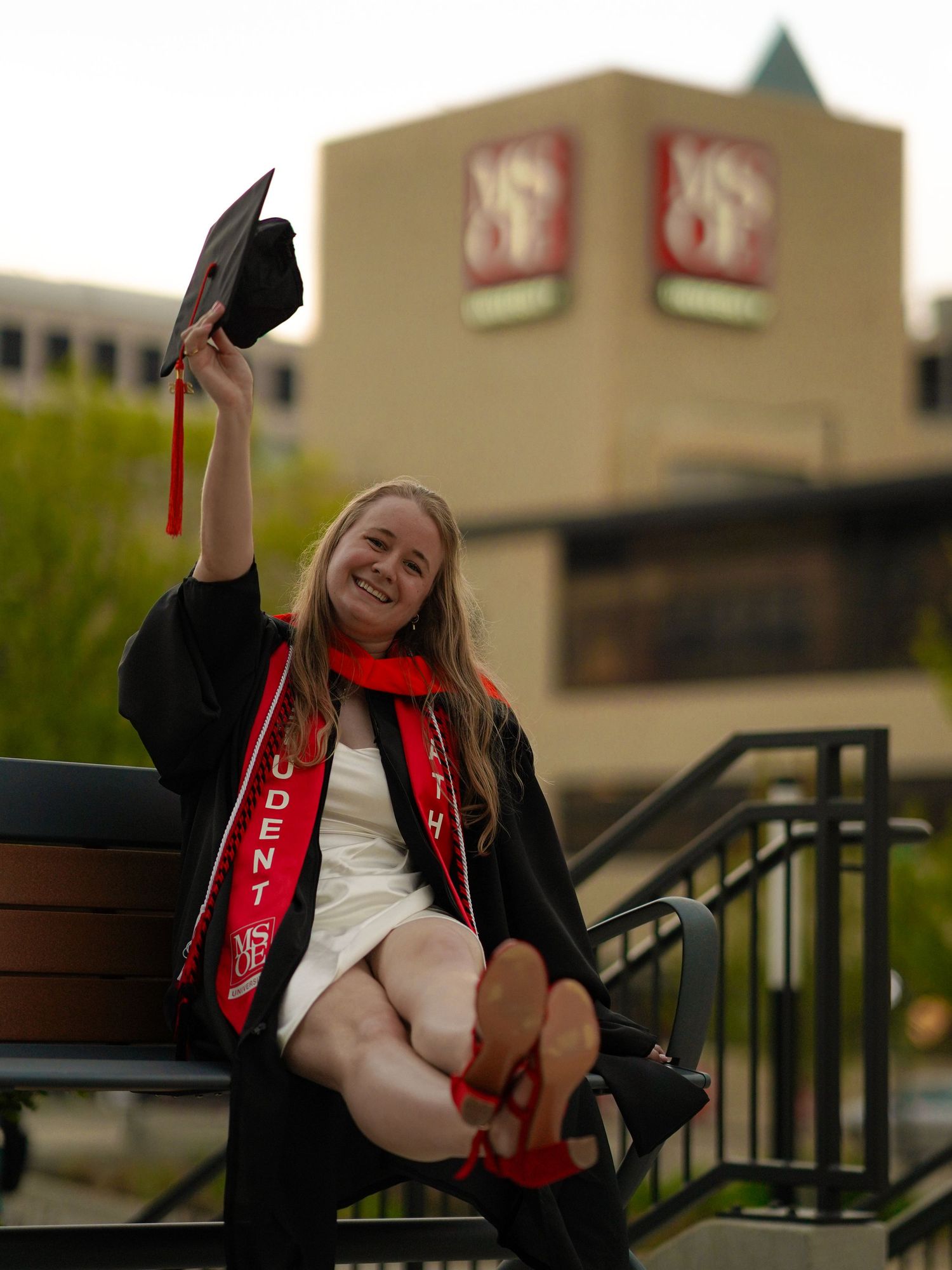 A graduate in black gown and red stole shows joyful expressions during outdoor graduation photo session.