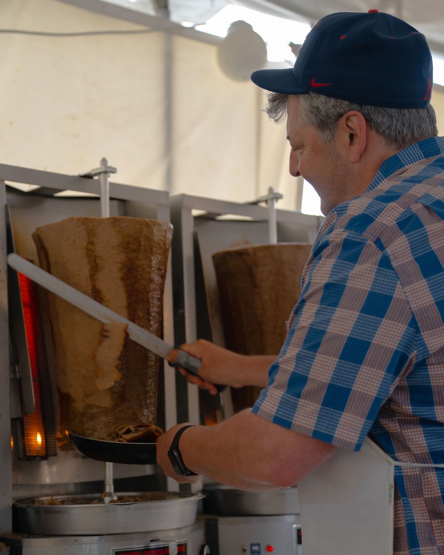 Person preparing food at a vertical rotisserie cooking station under a white tent.