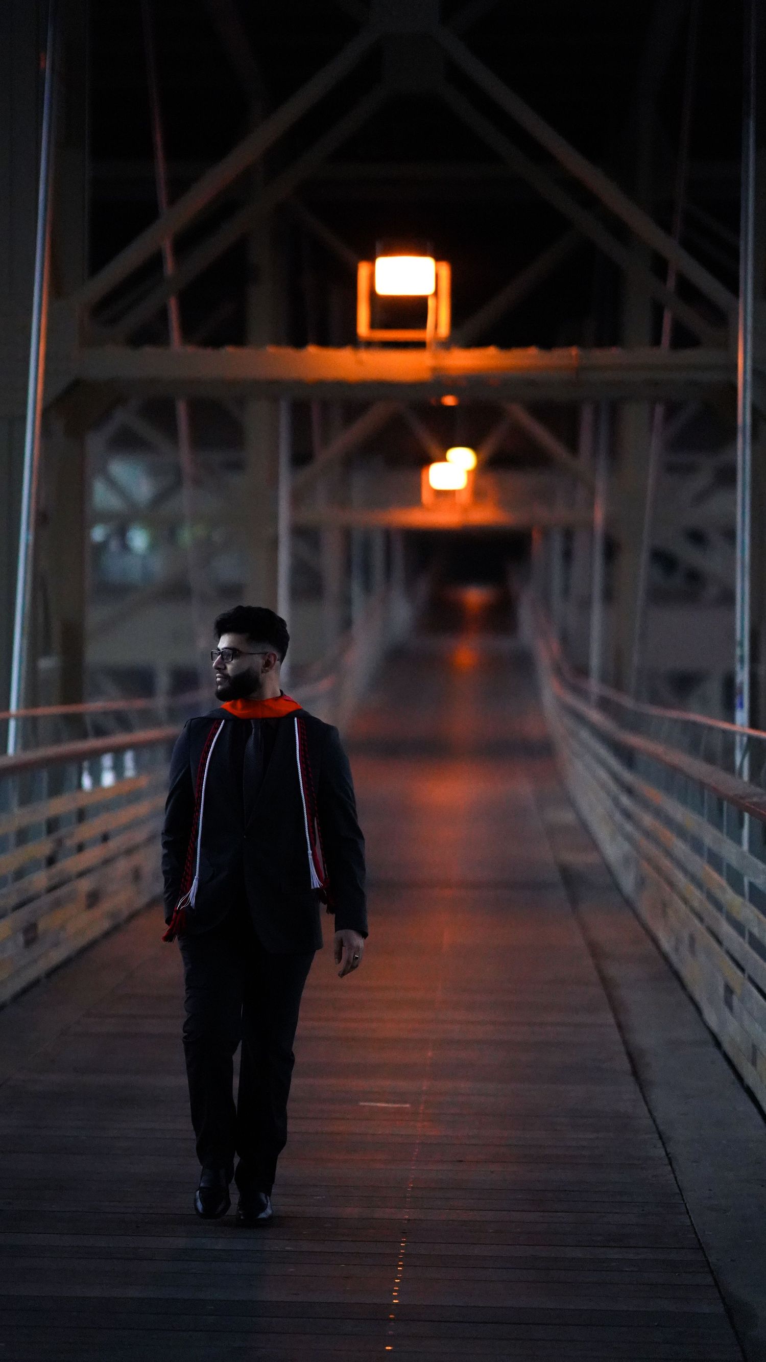Graduate walking alone on wooden bridge illuminated by warm overhead lights at dusk.