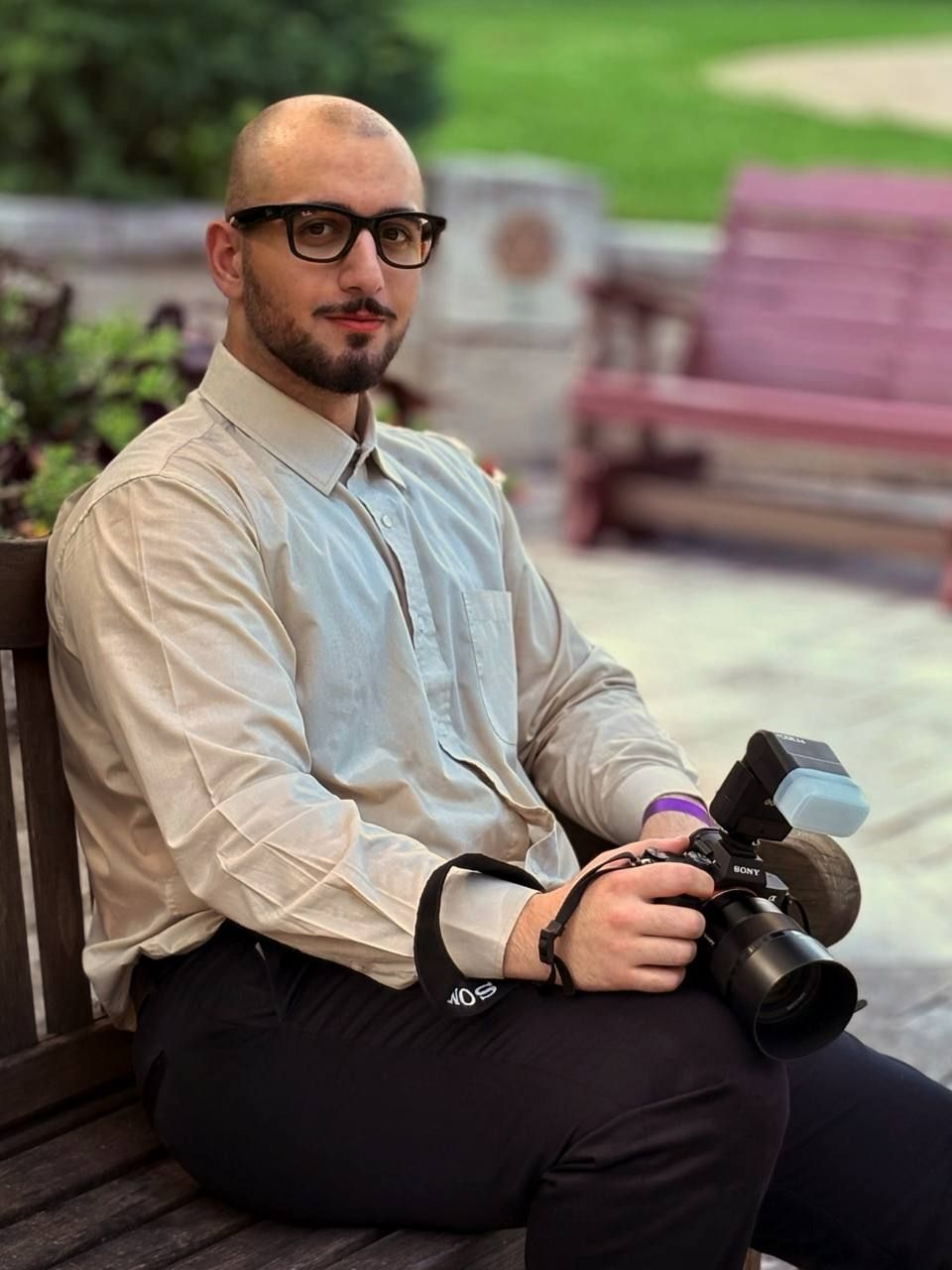 A person wearing glasses and business attire sits relaxed on a bench while holding a camera in an outdoor setting.