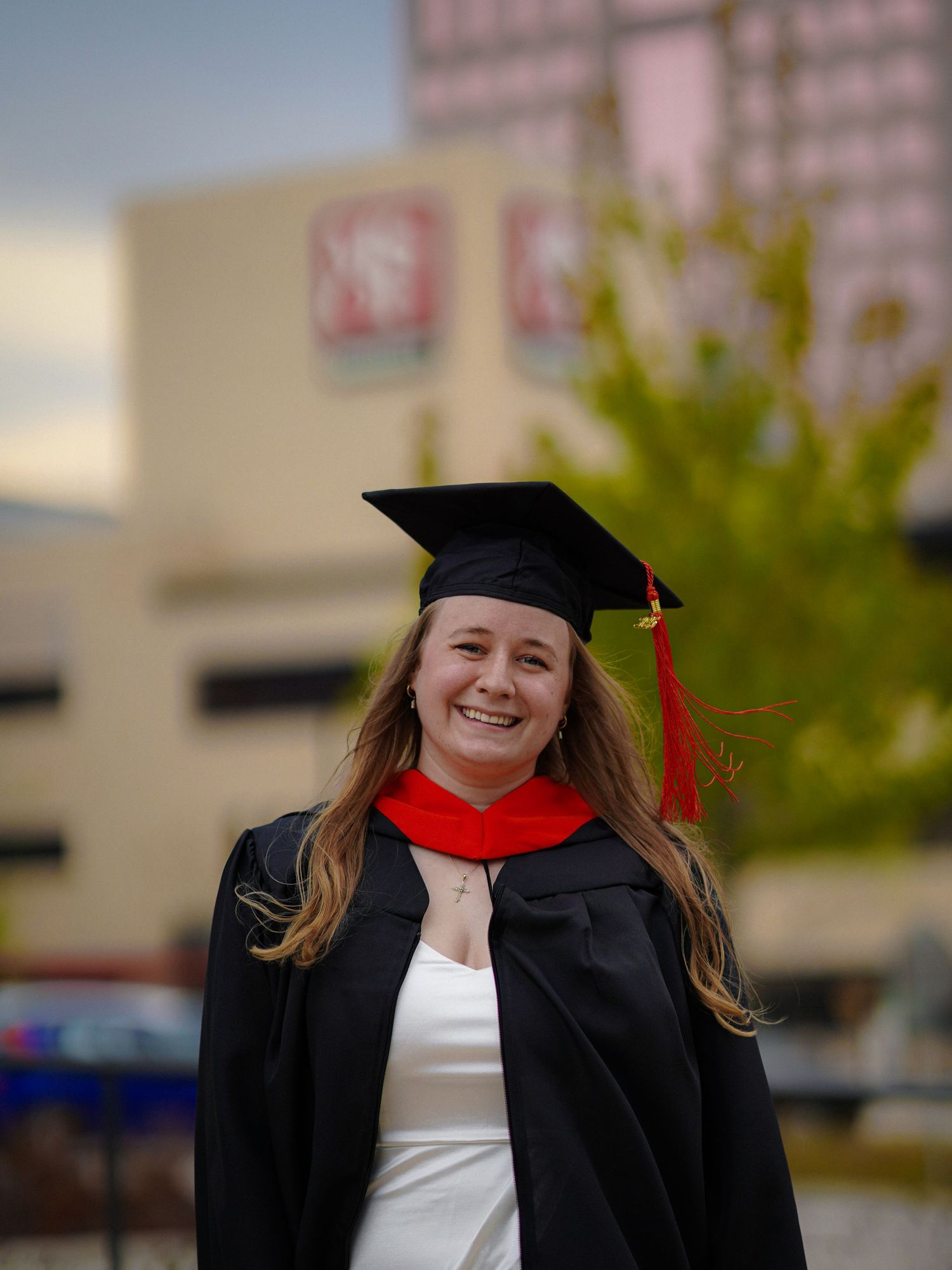 Graduate wearing traditional black cap and gown with red stole smiling outdoors on campus.