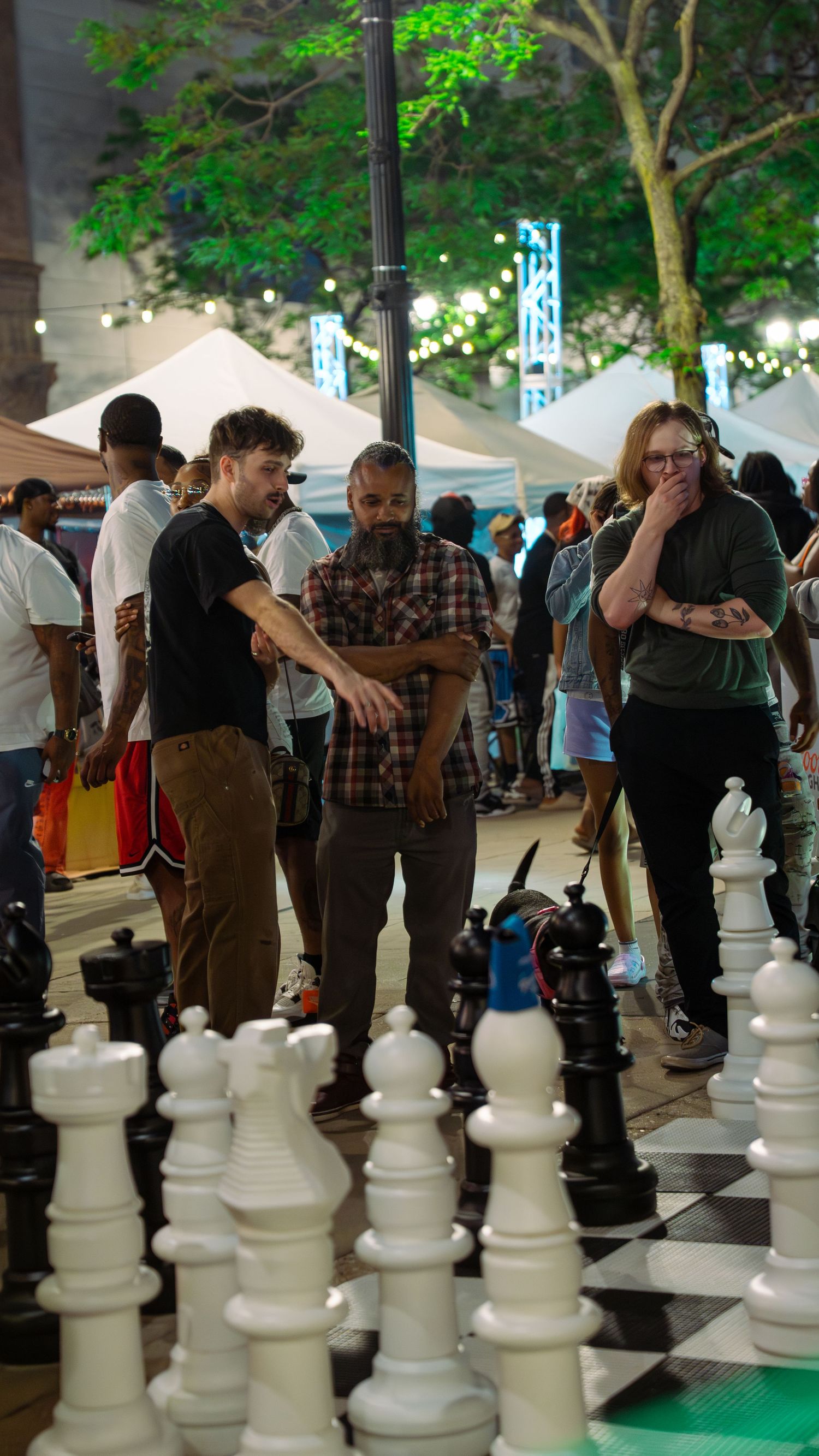 Crowd gathered around large outdoor chess game with illuminated trees in background.