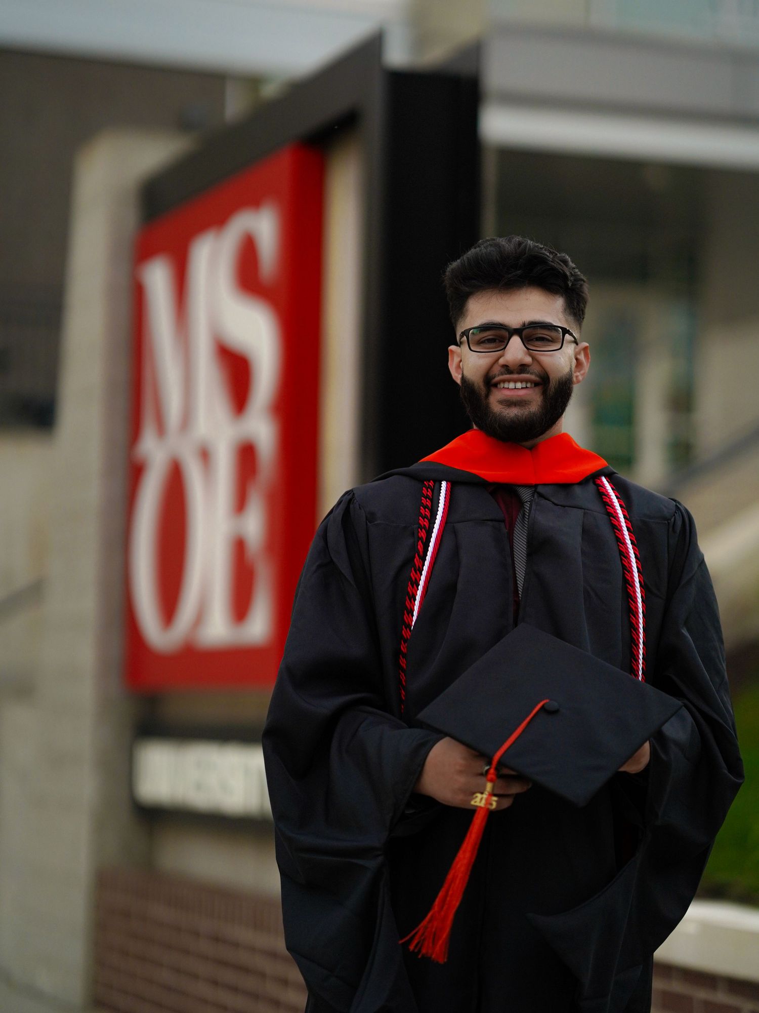 Graduate wearing black cap and gown with red stole celebrates commencement with bright smile.