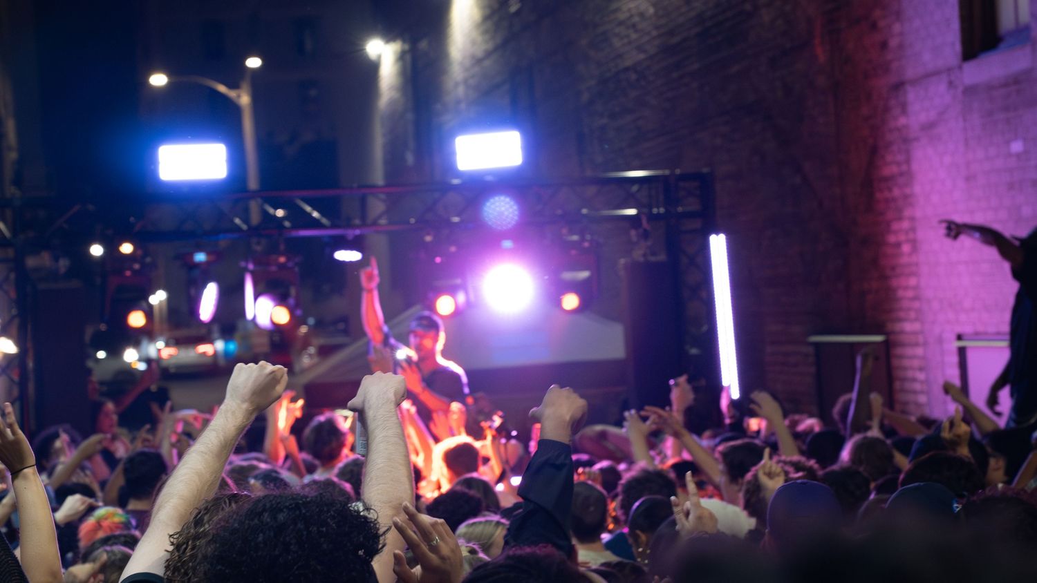 Concert crowd with raised hands under colorful stage lighting.