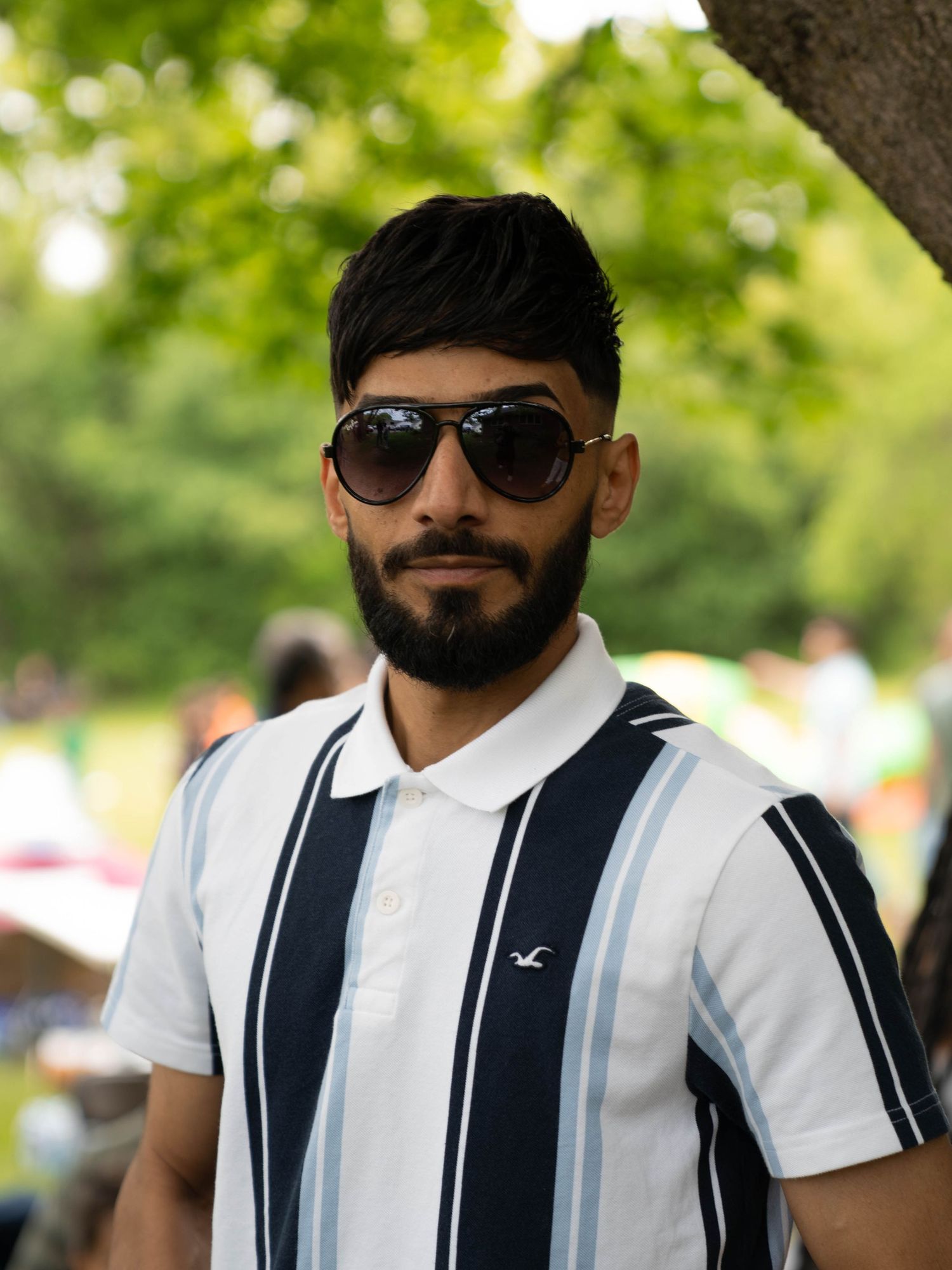 A person in sunglasses wearing a white and navy striped polo shirt stands outdoors against a blurred green background.