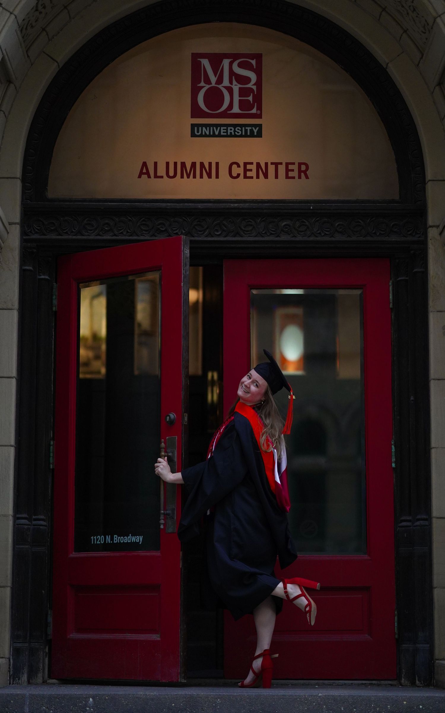 A person poses playfully in front of red doors at a university alumni center.