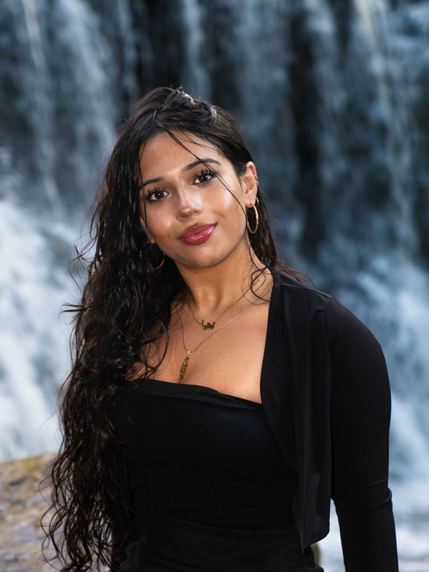 Woman with long dark hair wearing black attire poses in front of cascading waterfall backdrop.