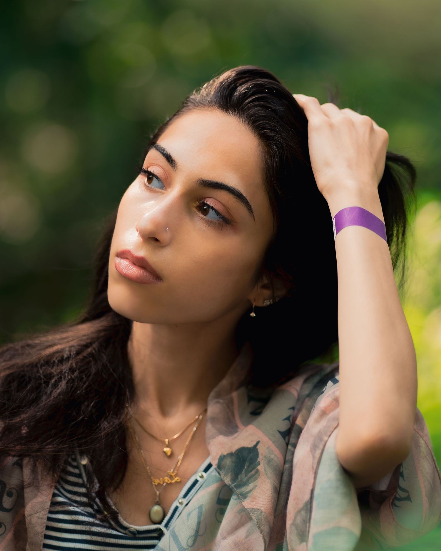 A portrait of someone wearing a floral jacket and purple wristband while fixing their long dark hair outdoors.