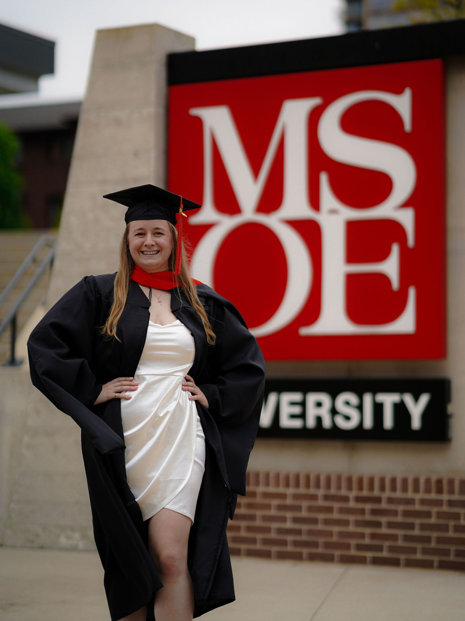 A graduate poses in traditional black cap and gown against red university signage during a commencement photo session.
