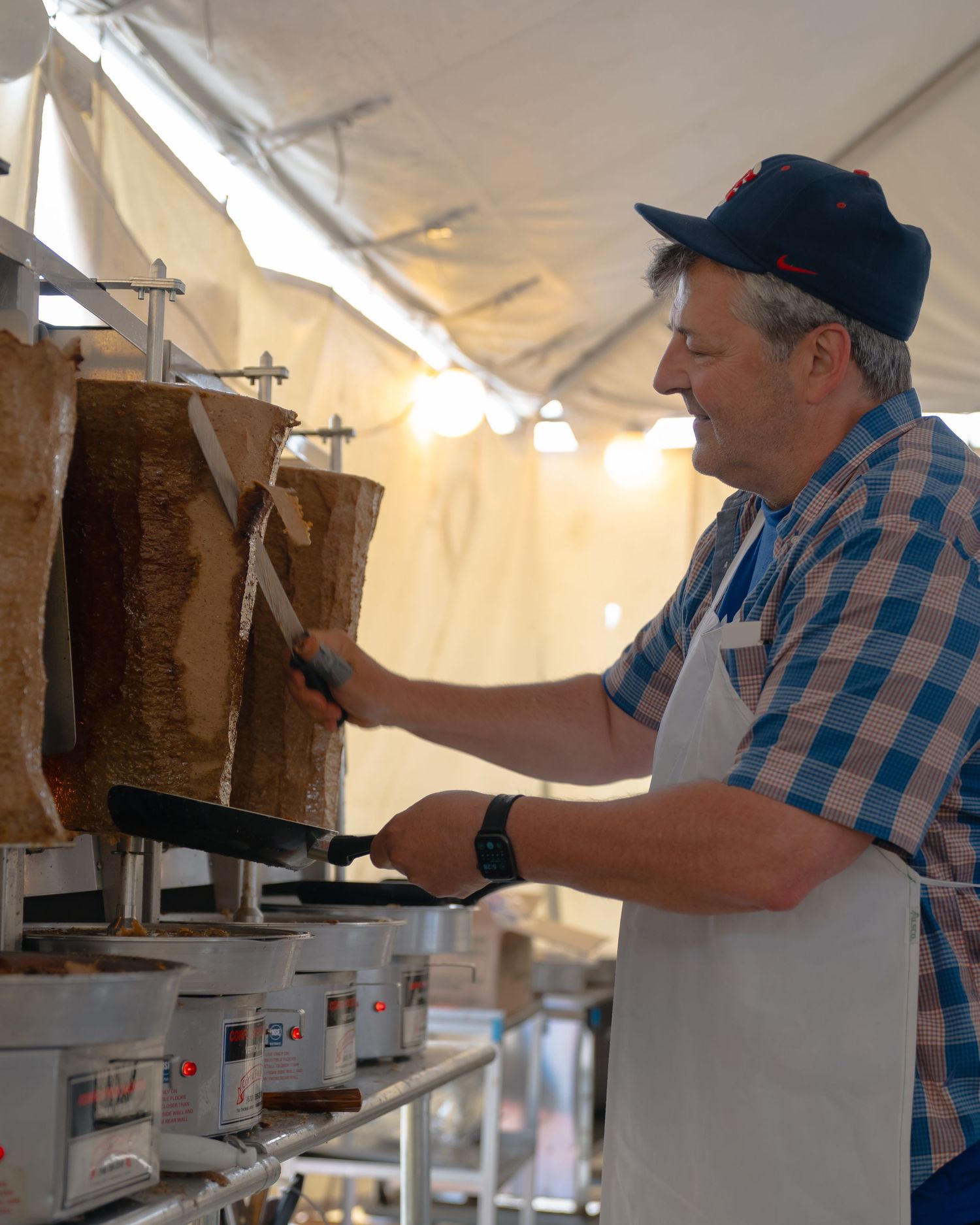 Cook in blue plaid shirt tending to vertical cooking equipment at an outdoor food station.