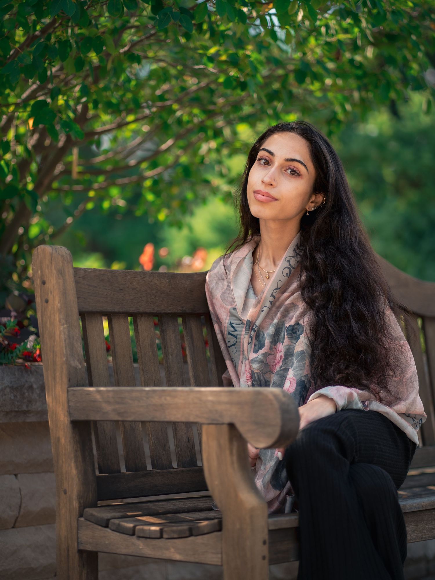 A person with long dark hair sits thoughtfully on a wooden bench under green foliage in a peaceful outdoor setting.