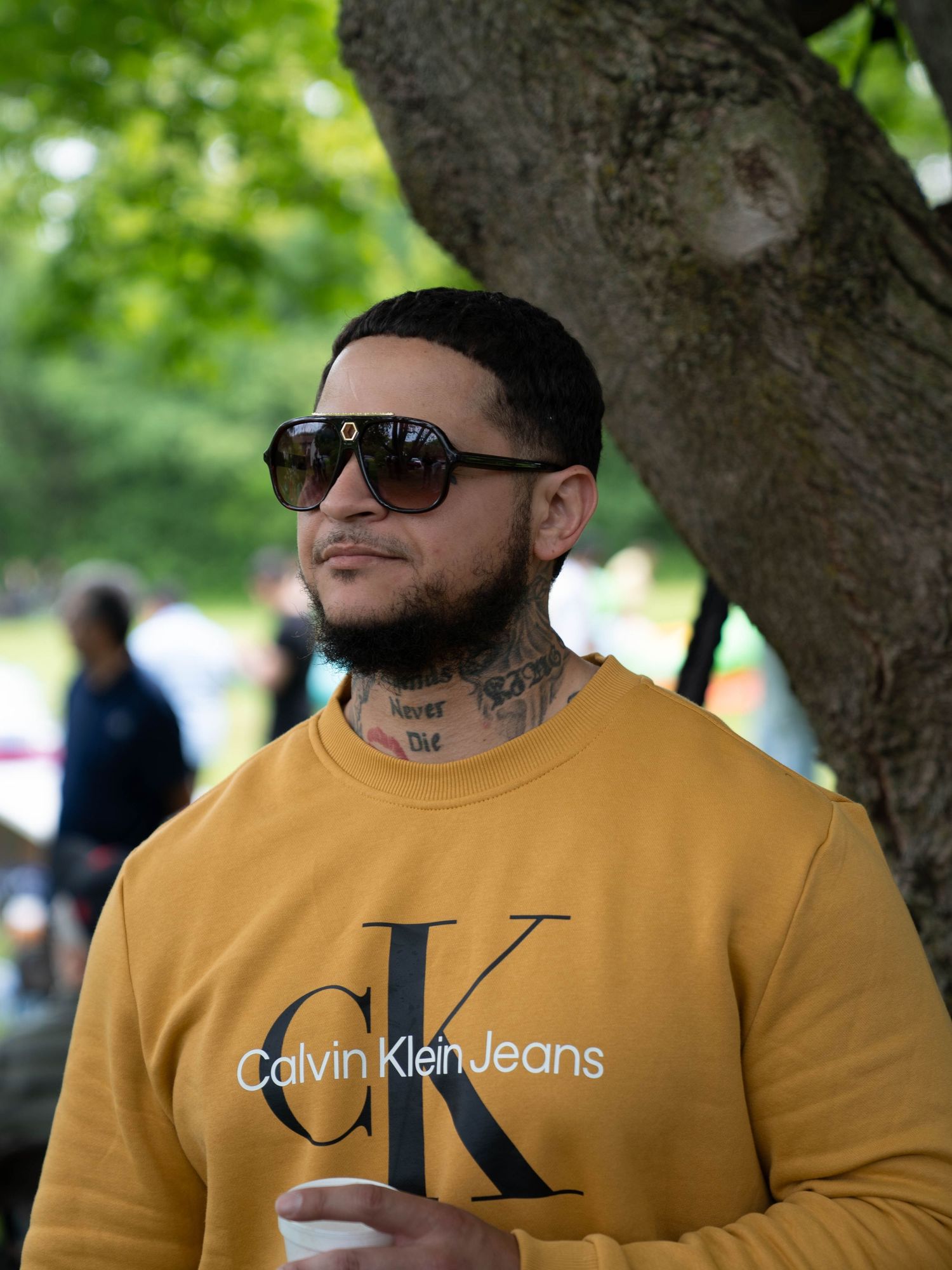 Person in sunglasses wearing a yellow Calvin Klein Jeans logo t-shirt standing near a tree in an outdoor setting.