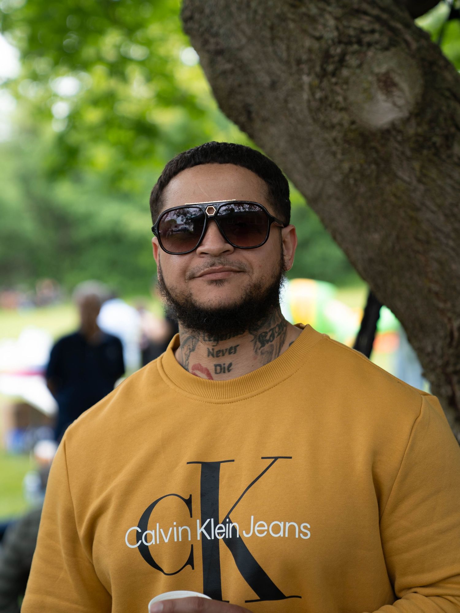 Person wearing yellow Calvin Klein sweatshirt and sunglasses posing outdoors under trees.