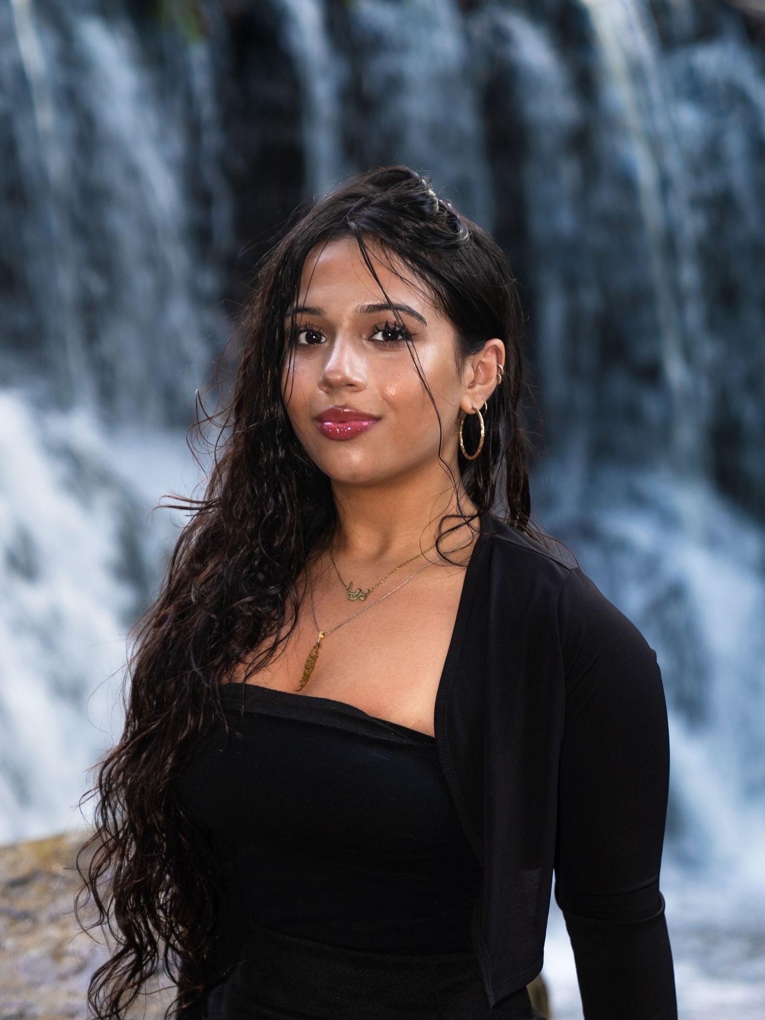 Woman with long dark hair wearing a black top poses in front of cascading waterfall backdrop.