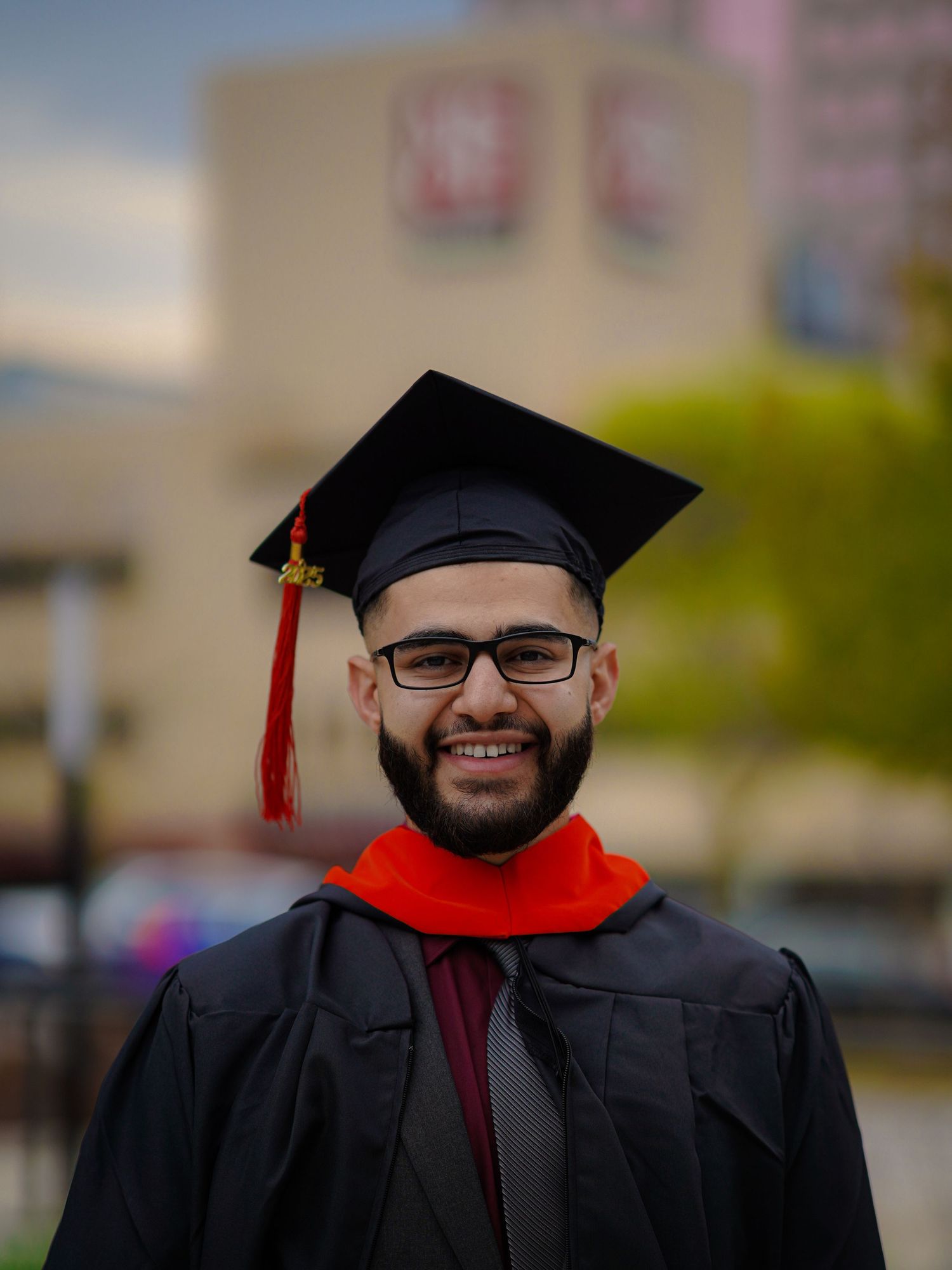 A graduate in academic regalia with black gown and red stole smiles outdoors during commencement ceremonies.