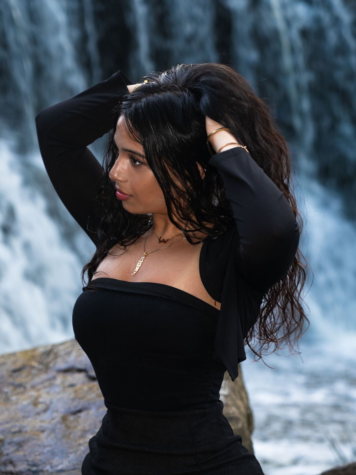 Dark-haired woman in black long-sleeve dress poses near a cascading waterfall in a dramatic outdoor portrait setting.