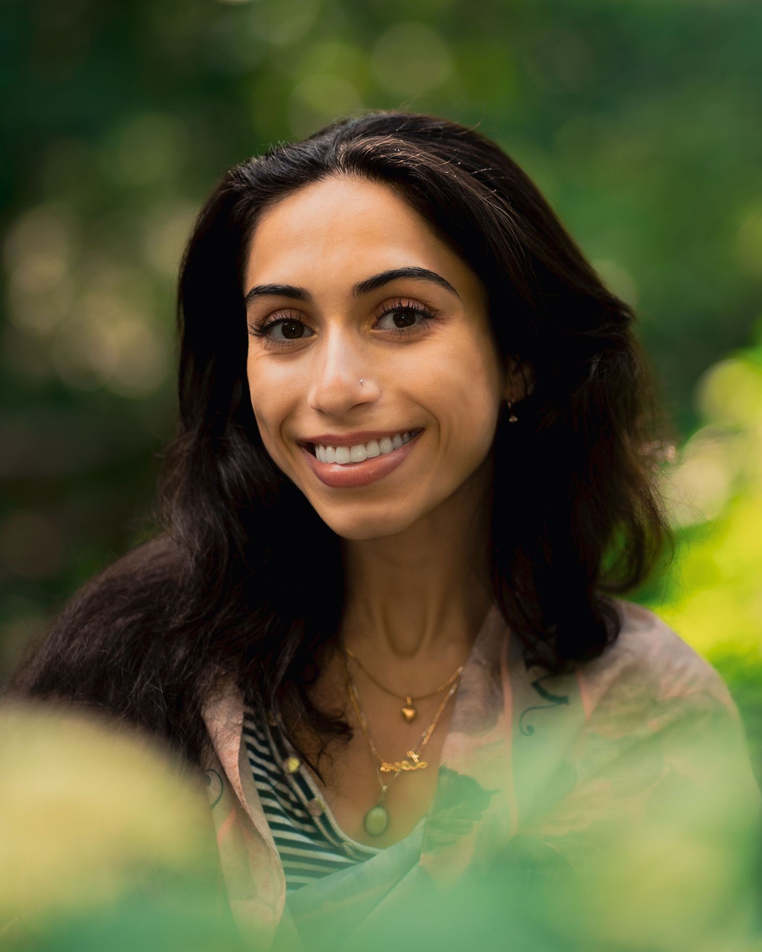 Smiling person with long dark hair wearing a necklace poses outdoors against a blurred green natural background.