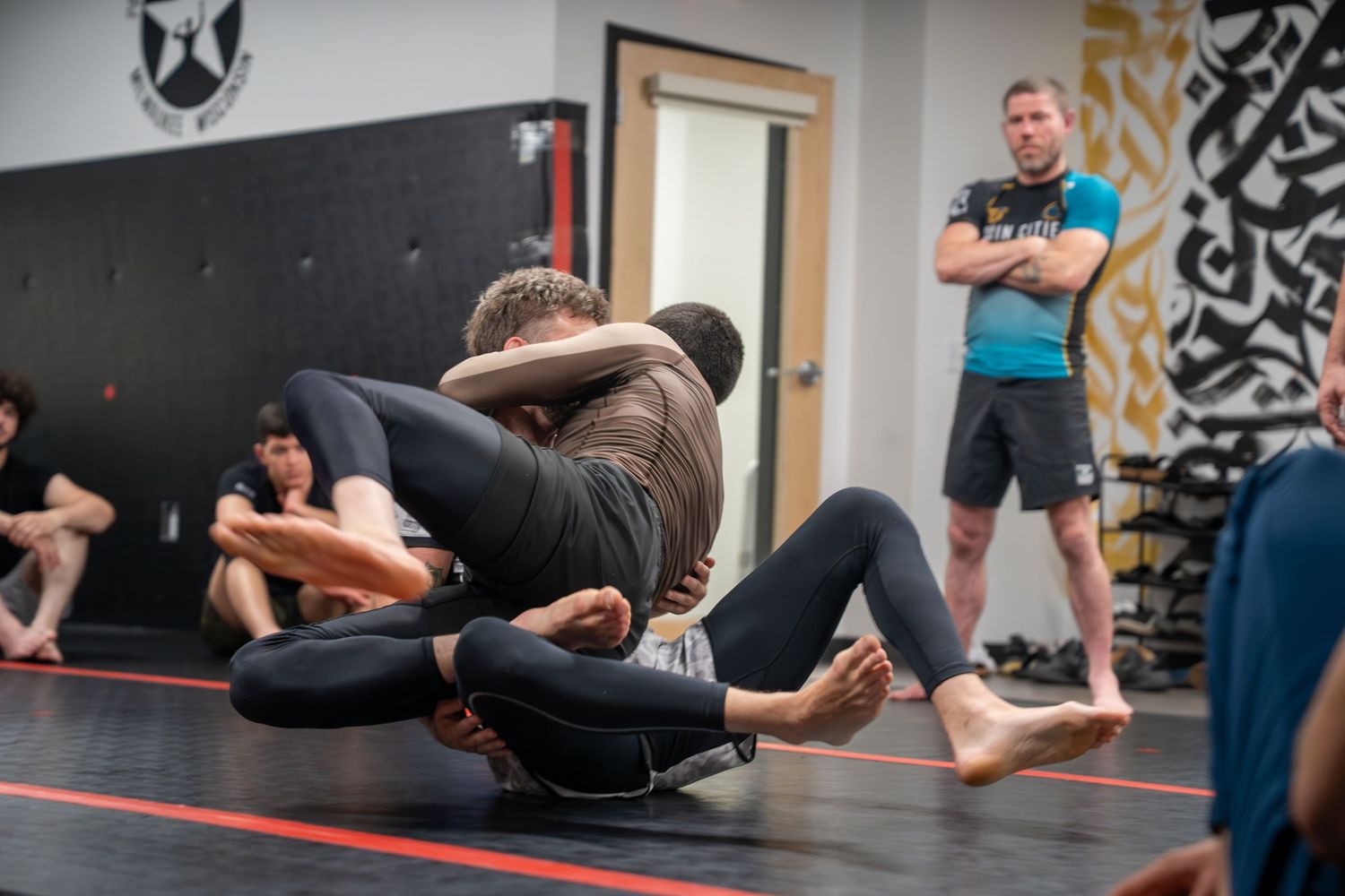 Athletes practicing grappling moves on black mats in a martial arts gym.