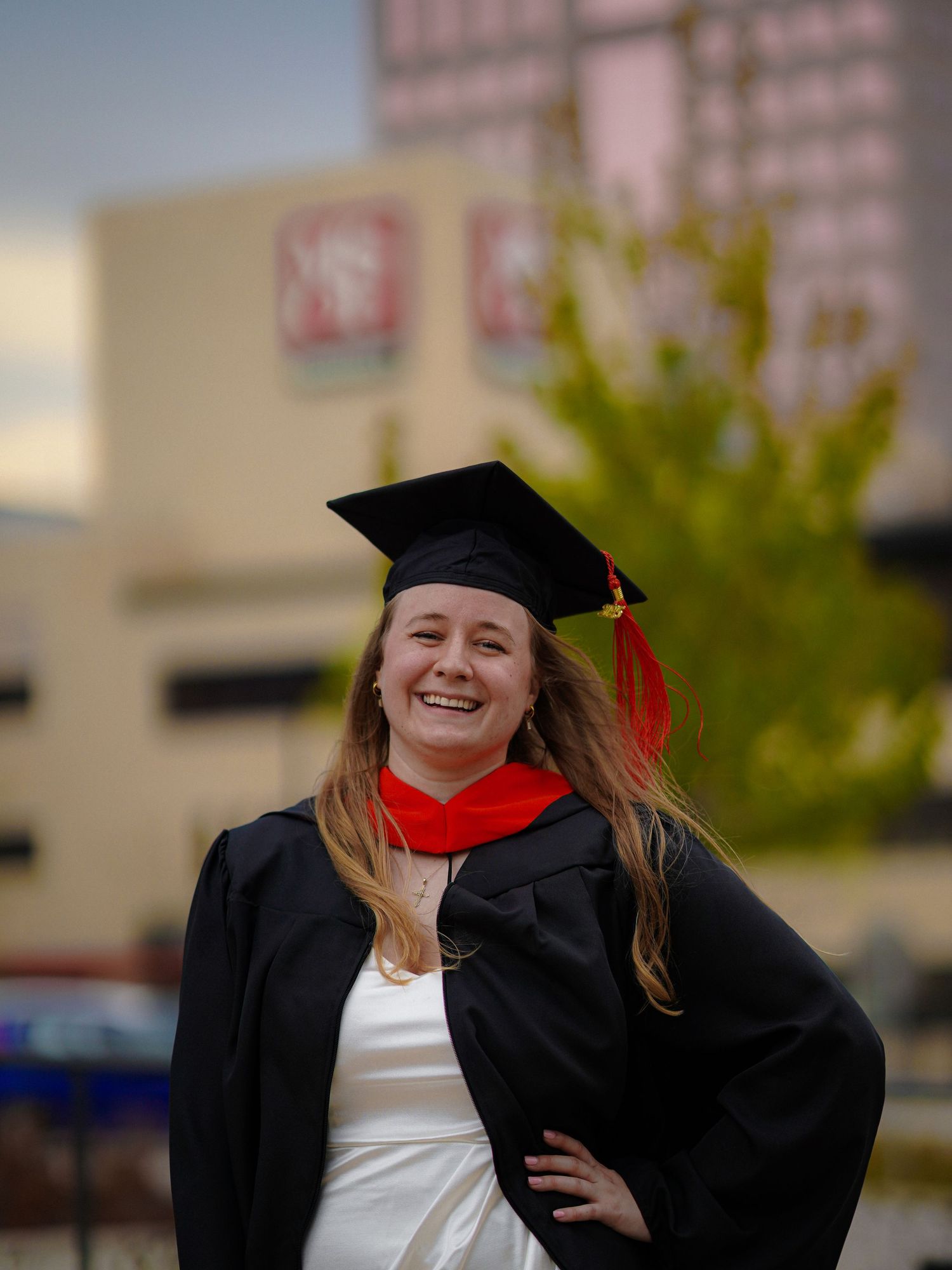 Graduate in black cap and gown with red stole poses confidently outdoors during commencement celebration.