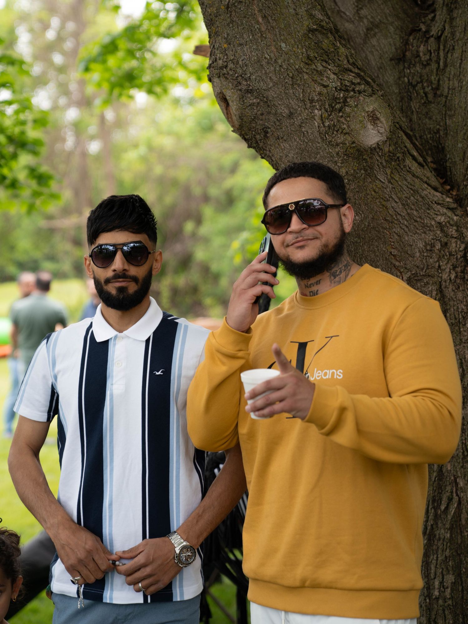Two people in sunglasses stand by a tree, with one wearing a striped shirt and another in a mustard yellow sweatshirt.