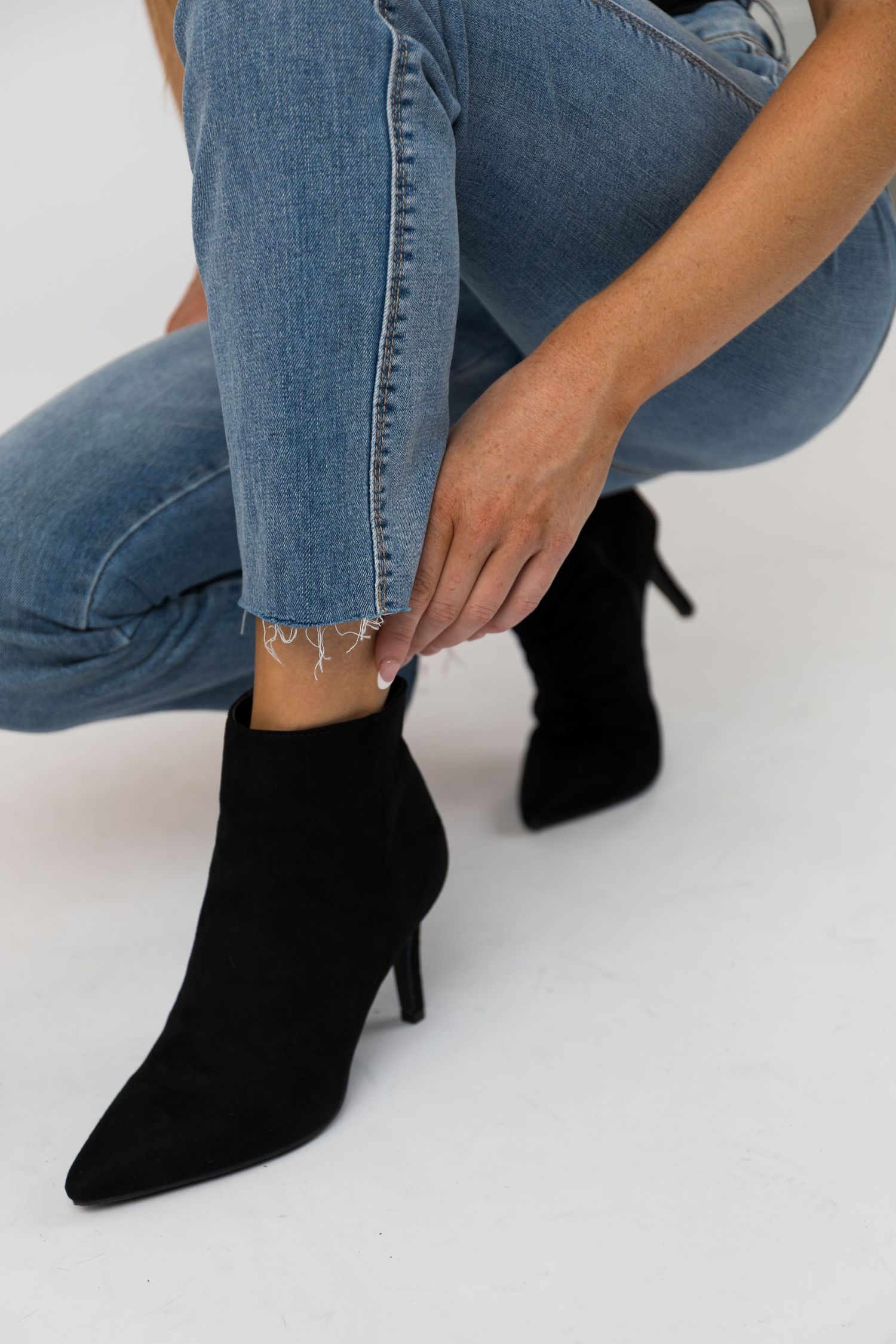 Black suede ankle boots with stiletto heels styled with blue jeans against a white background.