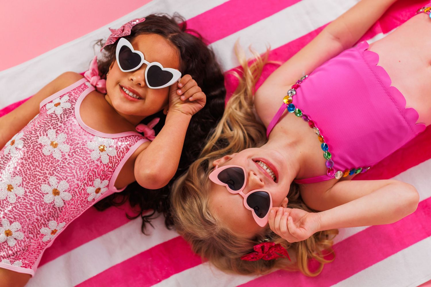 Two people wearing heart-shaped sunglasses lay on pink and white striped fabric while smiling and posing playfully.