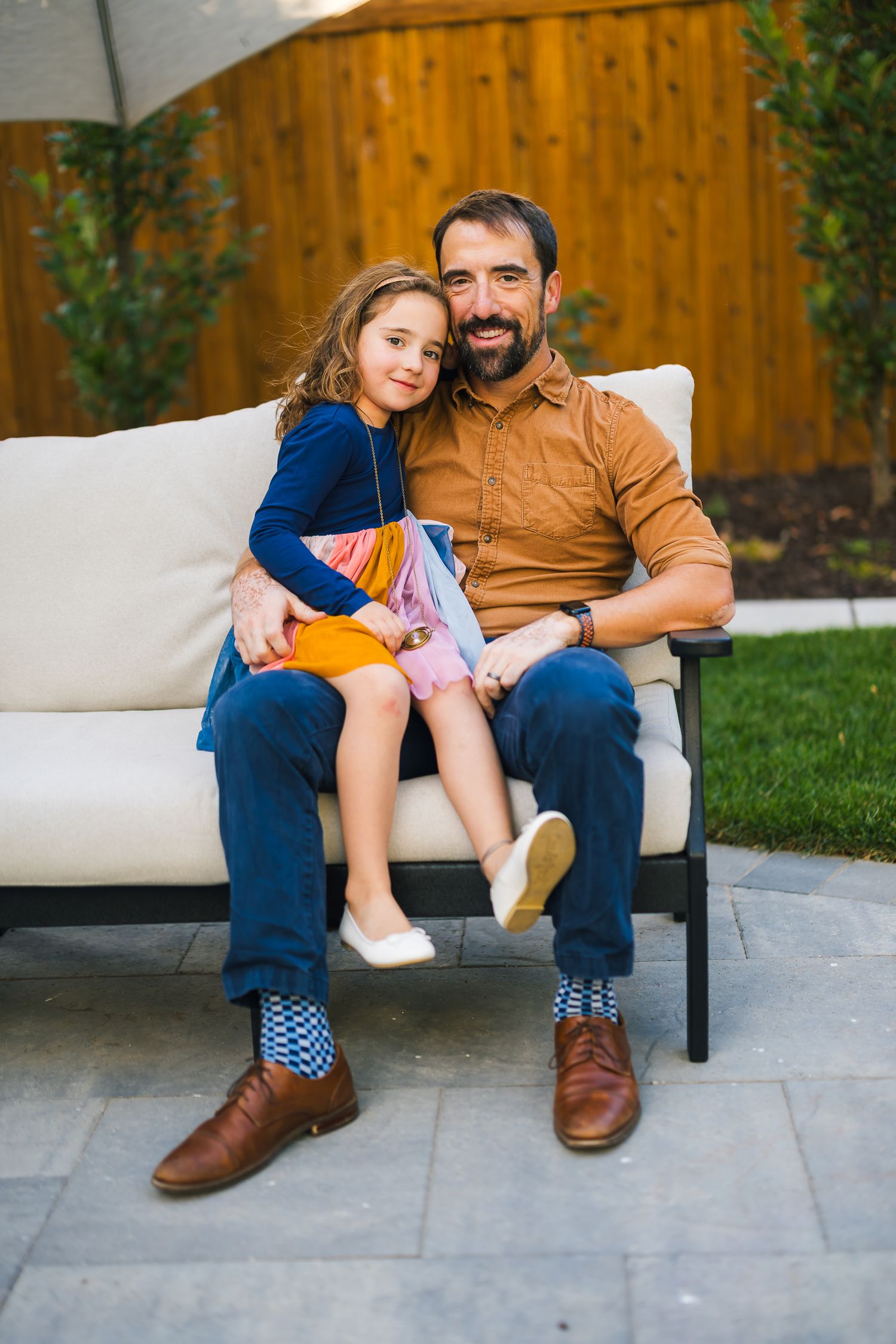Parent and child share a tender moment on white outdoor furniture with colorful clothing against wooden fence.