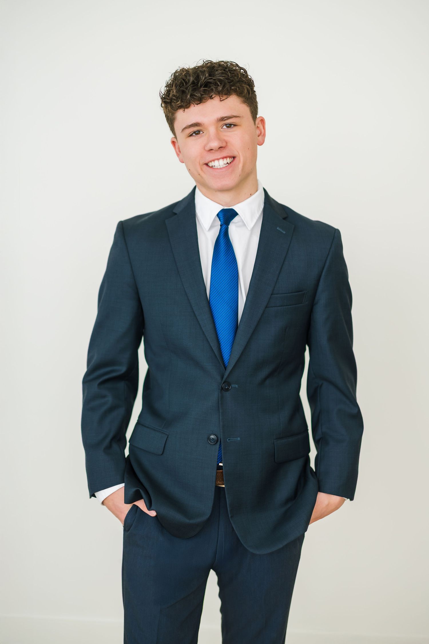 Young man in navy suit with blue tie smiling confidently against white background.