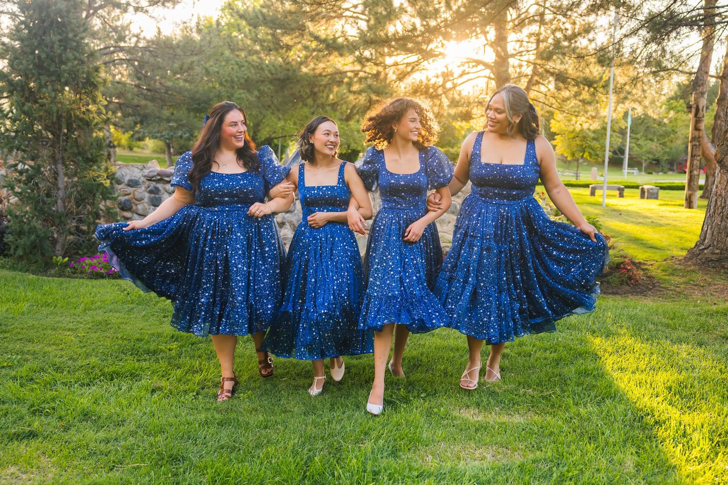 Four people in matching blue polka dot dresses twirl together on a sunlit lawn at sunset.