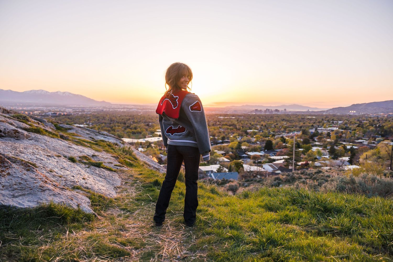 Person in casual attire stands on hilltop overlooking city during sunset.