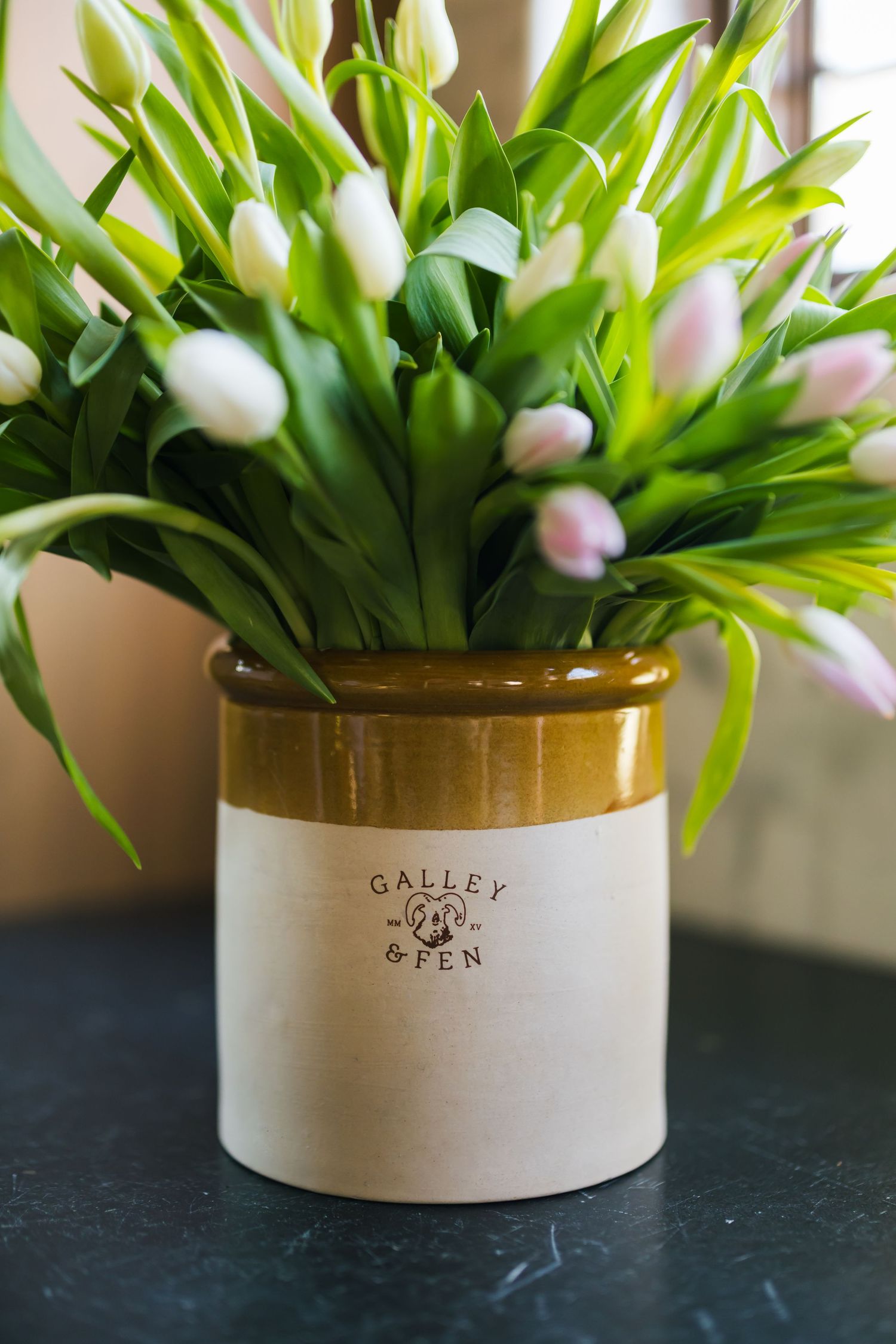 A ceramic Galley crock vase filled with fresh white and pink tulips sits on a dark surface.
