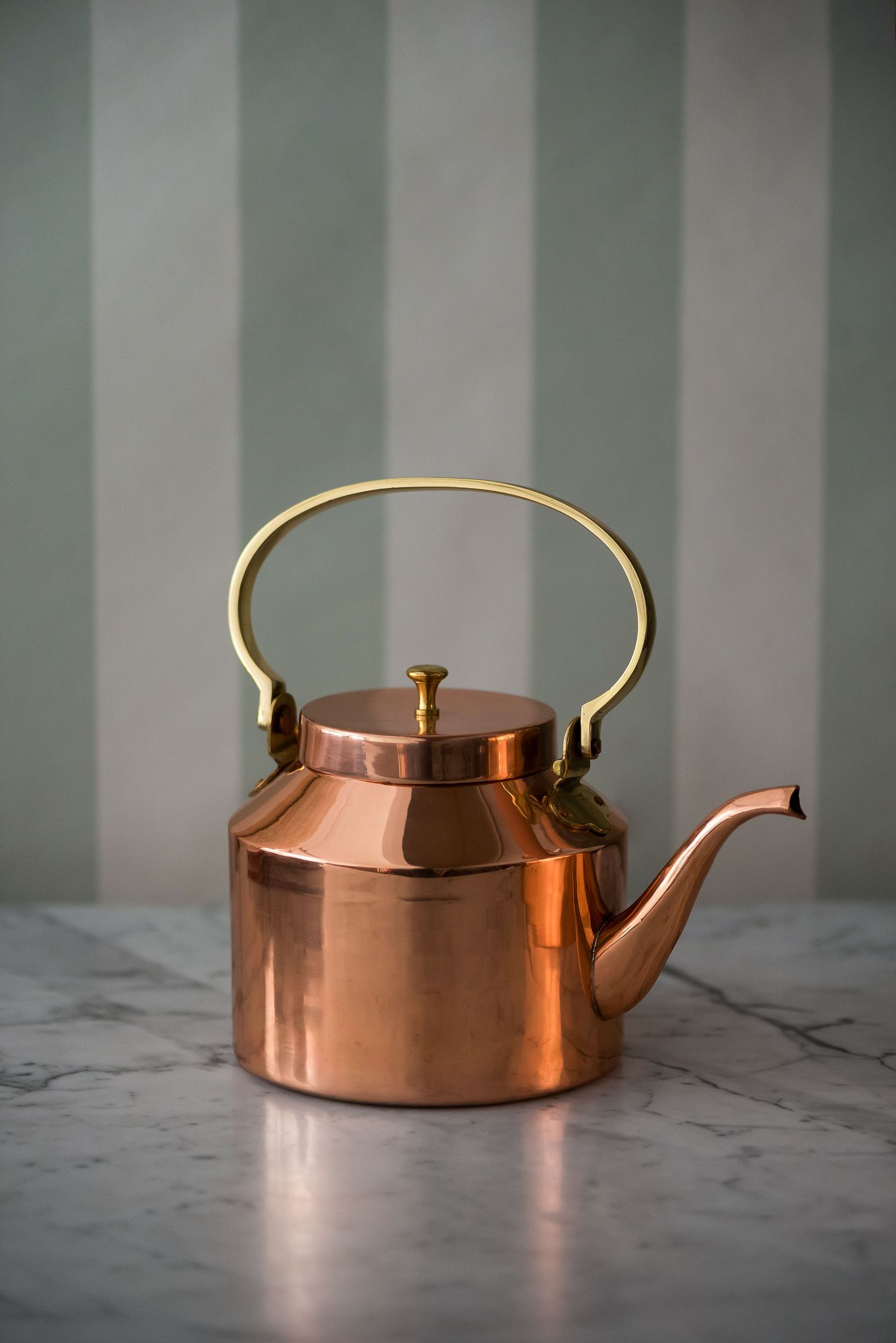 Copper teapot with brass handle on marble surface against striped wall background.