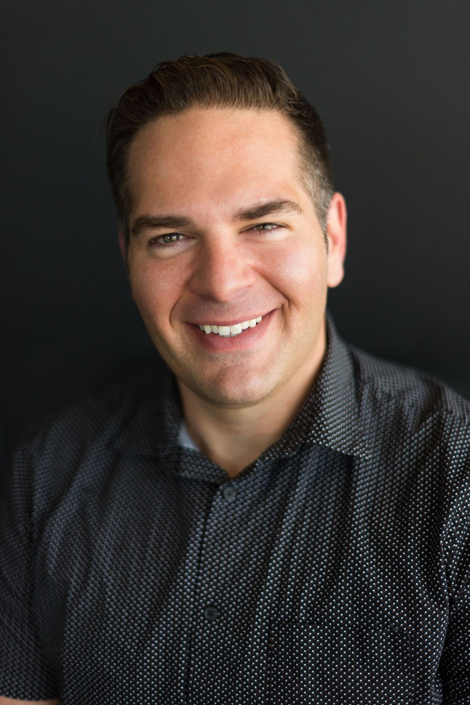 Professional headshot of a person in dark patterned shirt against black background.