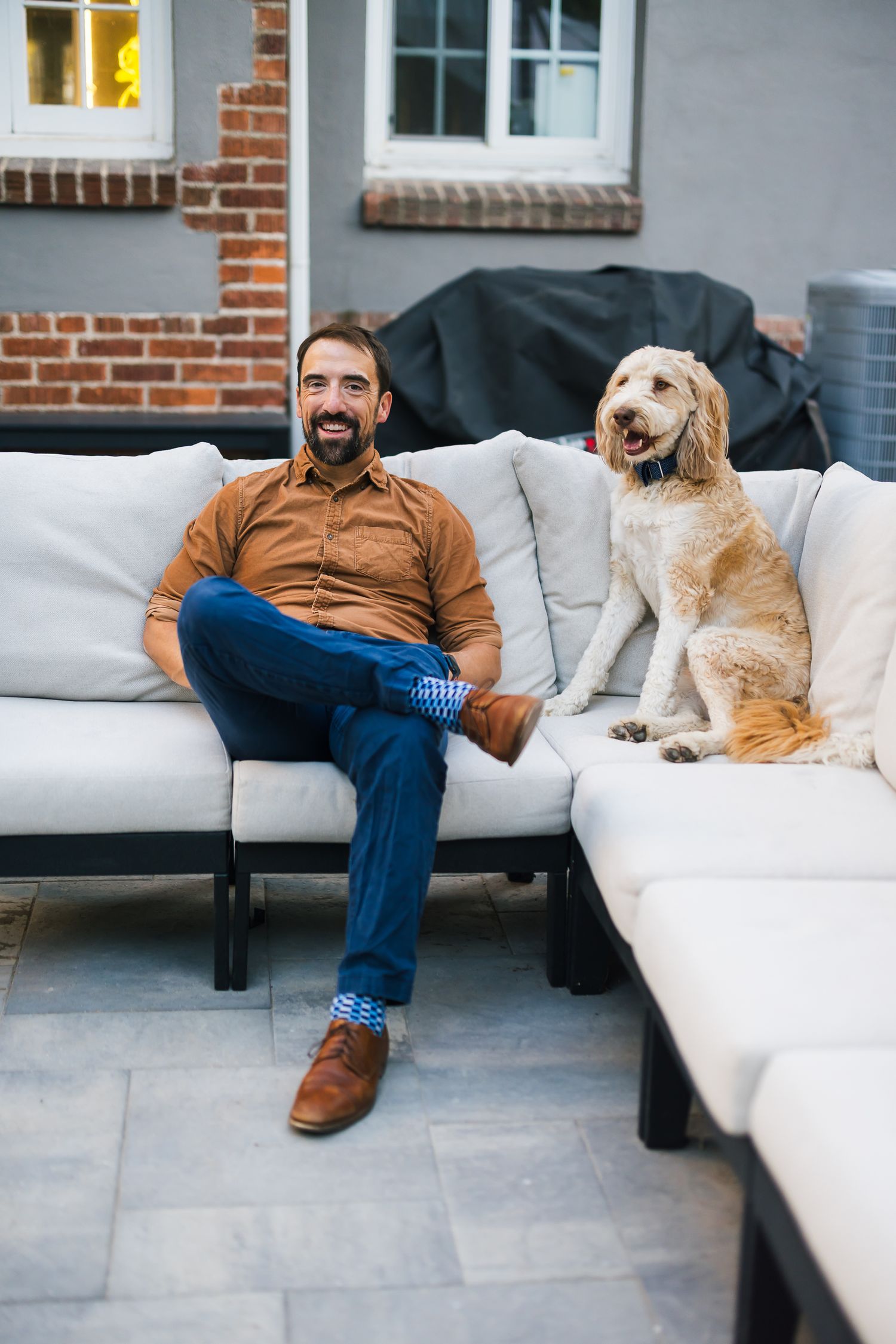 Man in brown shirt relaxes on outdoor patio furniture with golden retriever.