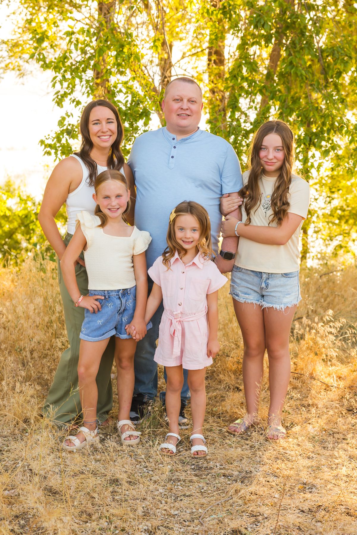 Family group portrait taken outdoors against a backdrop of sunlit trees and dry grass.