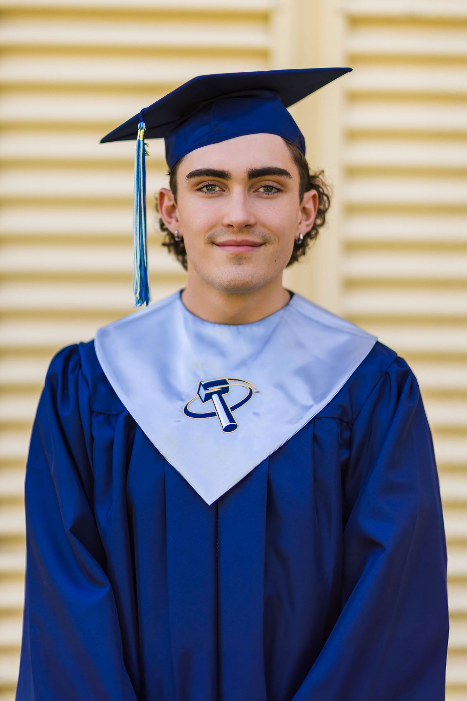 Young man in blue graduation cap and gown with light blue sash smiling for senior portrait photo.