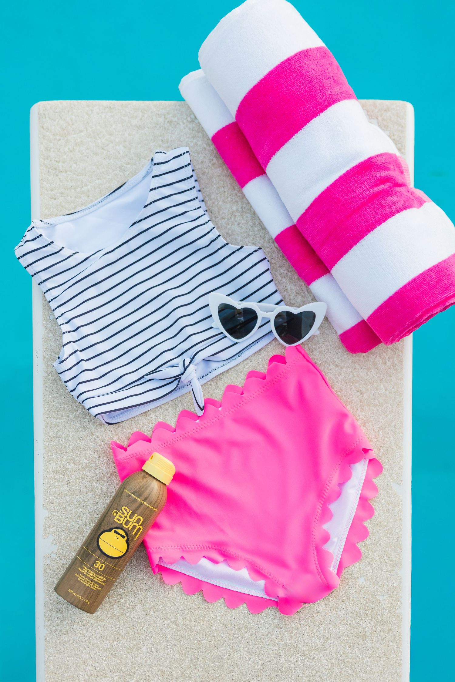 Striped navy and white bikini top with hot pink bottoms and tanning oil laid on sandy beach.
