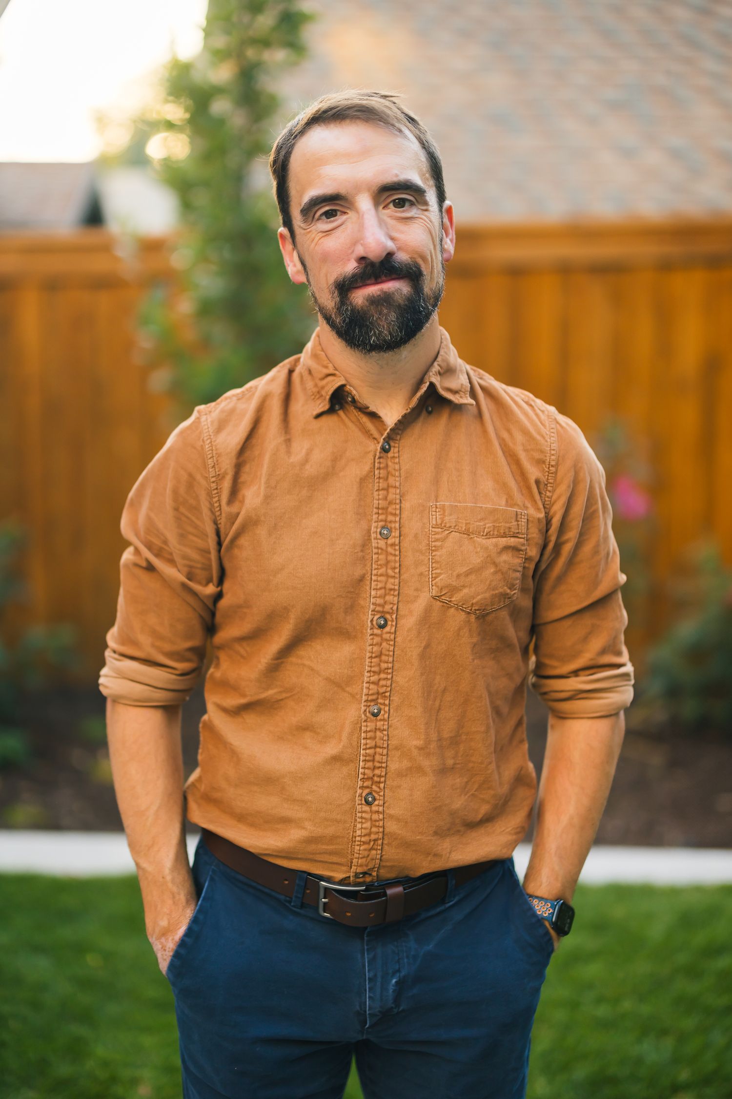 Casual portrait of person in brown button-down shirt against wooden fence backdrop.