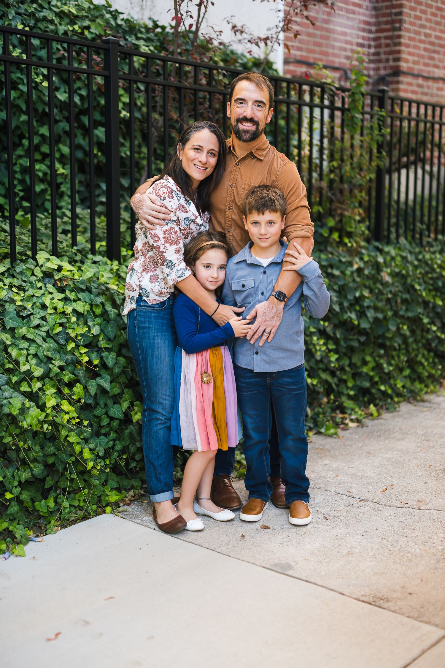 Family of four poses together against iron fence and lush green garden backdrop.